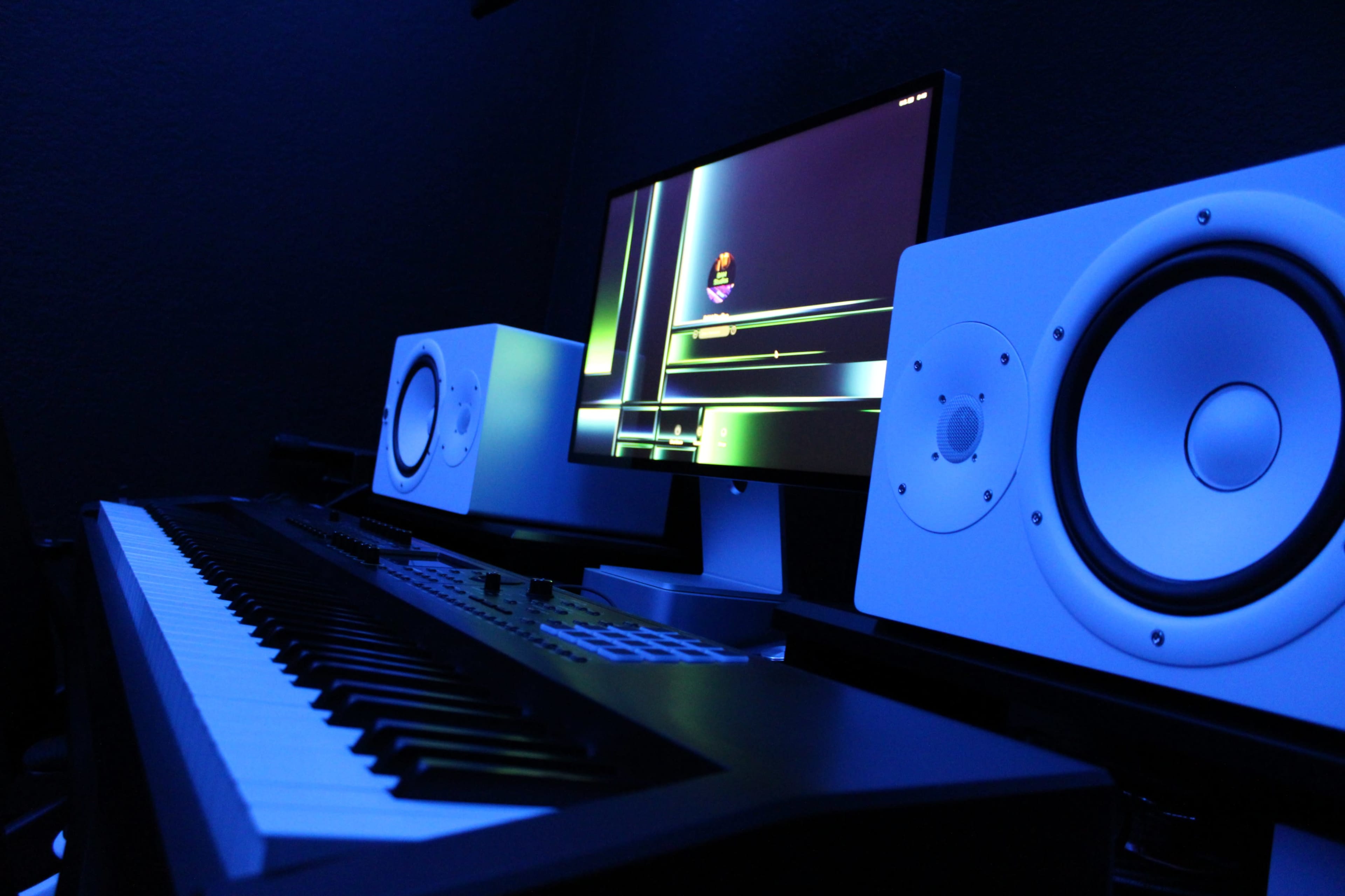 A digital music studio setup features a keyboard, a computer monitor displaying a graphical interface, and two white studio monitors under blue lighting.