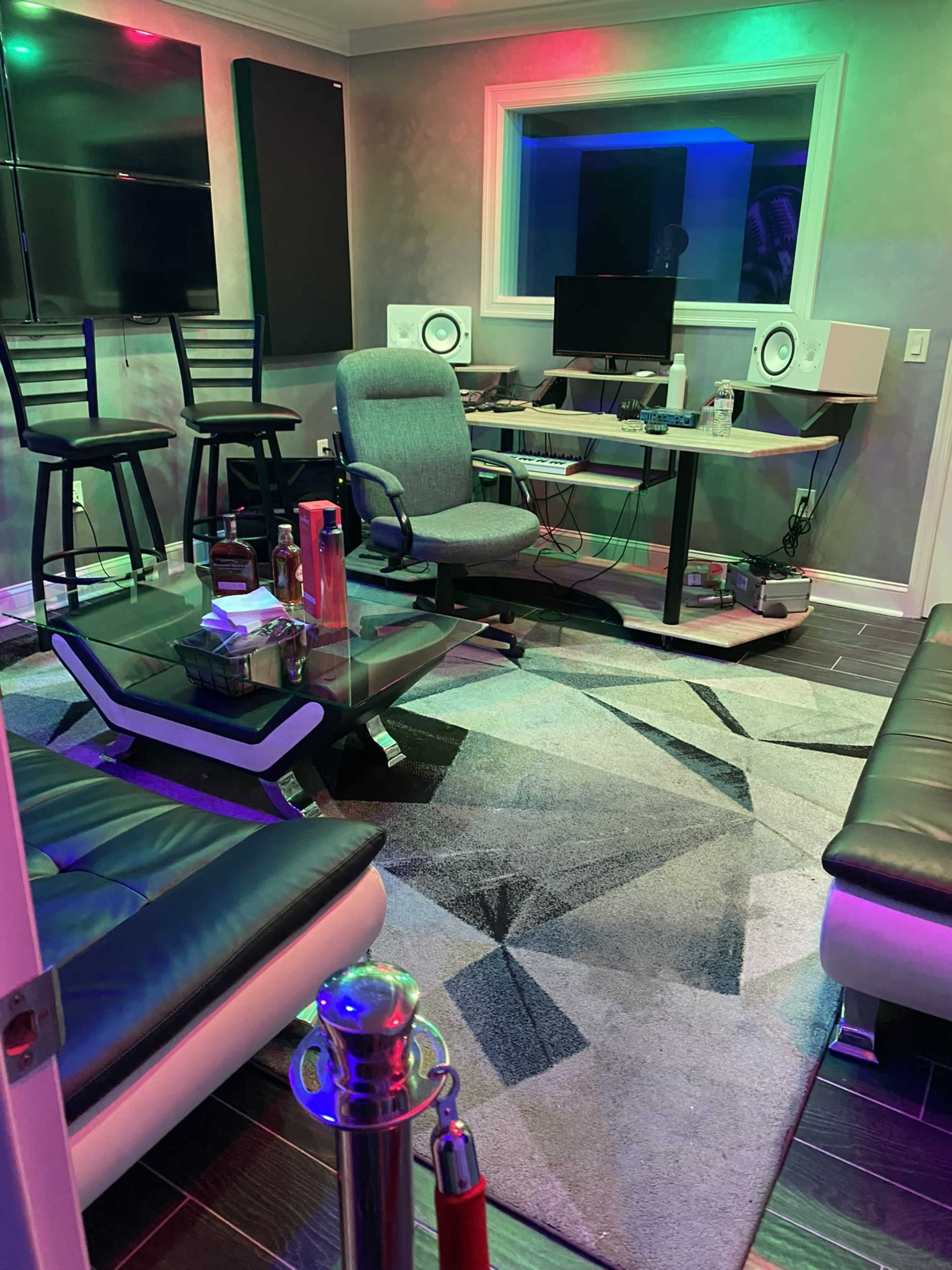 The image shows a modern recording studio with a desk, audio equipment, two speakers, and seating arrangements.