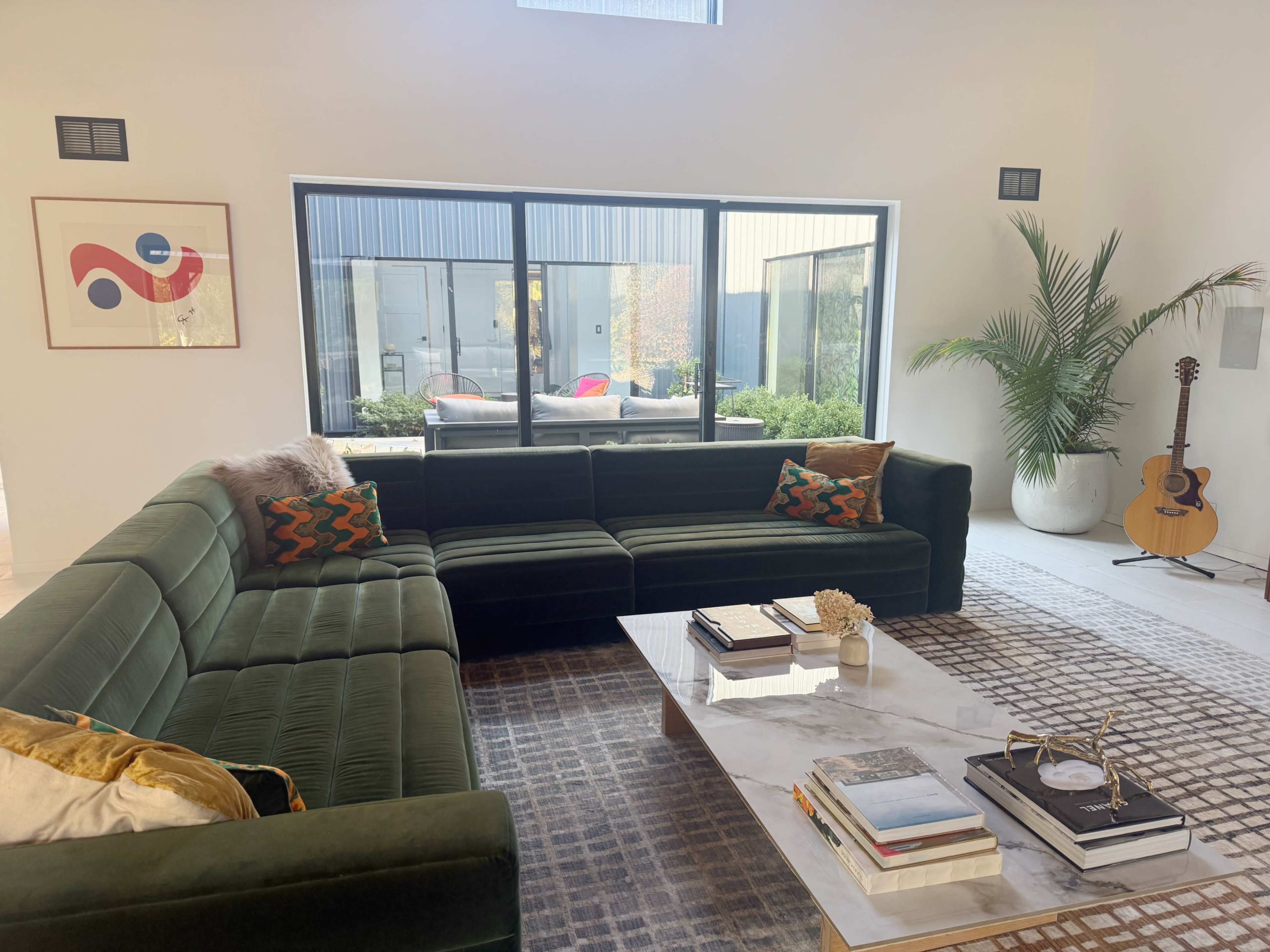 A modern living room features a dark green sectional sofa, a marble coffee table, and a large window overlooking an outdoor space with a plant in the corner.