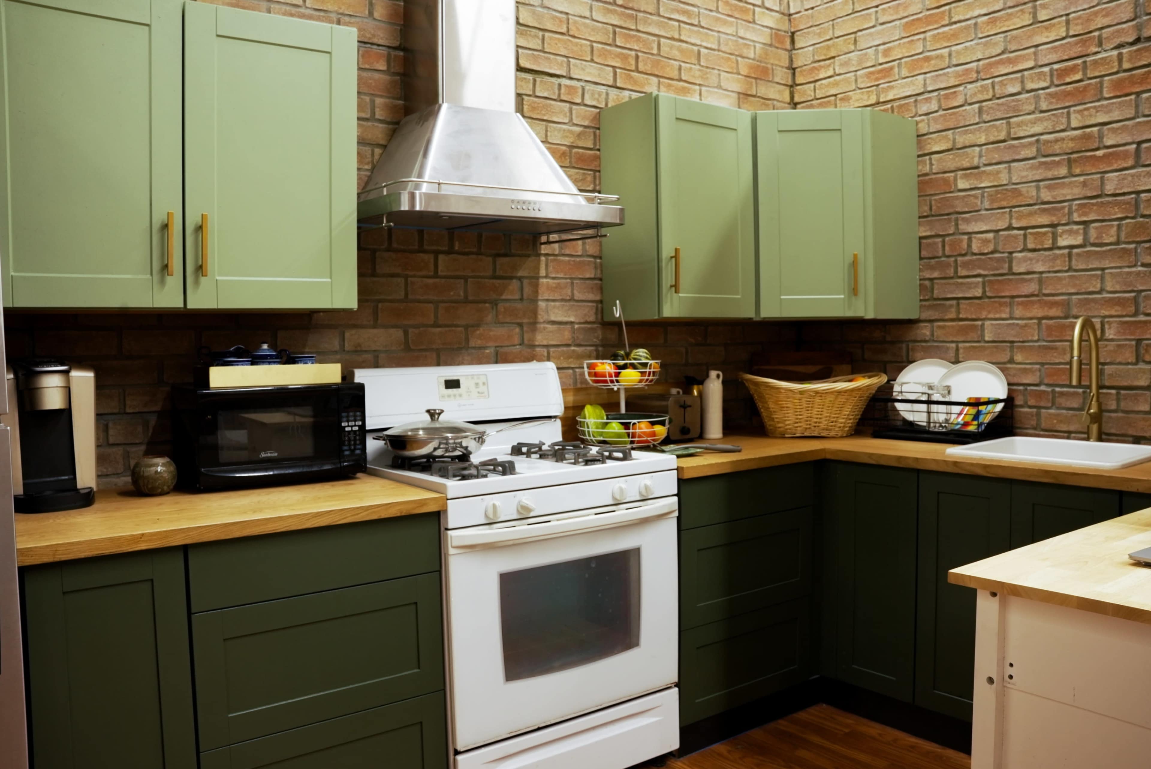 The image shows a kitchen featuring green cabinets, a white stove, a stainless steel range hood, and wooden countertops with various kitchen appliances and a basket.