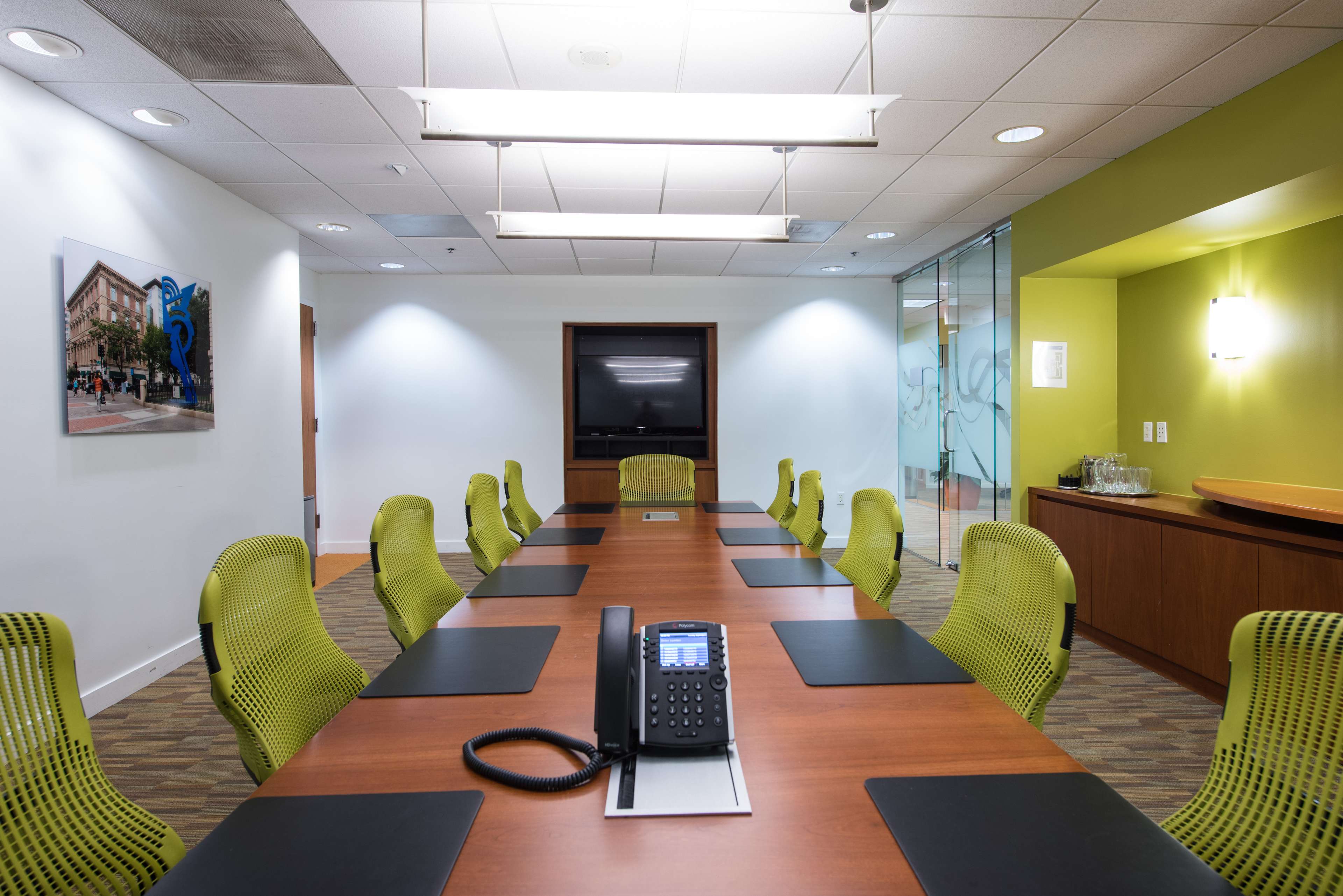 A conference room features a long wooden table with green chairs, a telephone at the center, and a television mounted on the wall.
