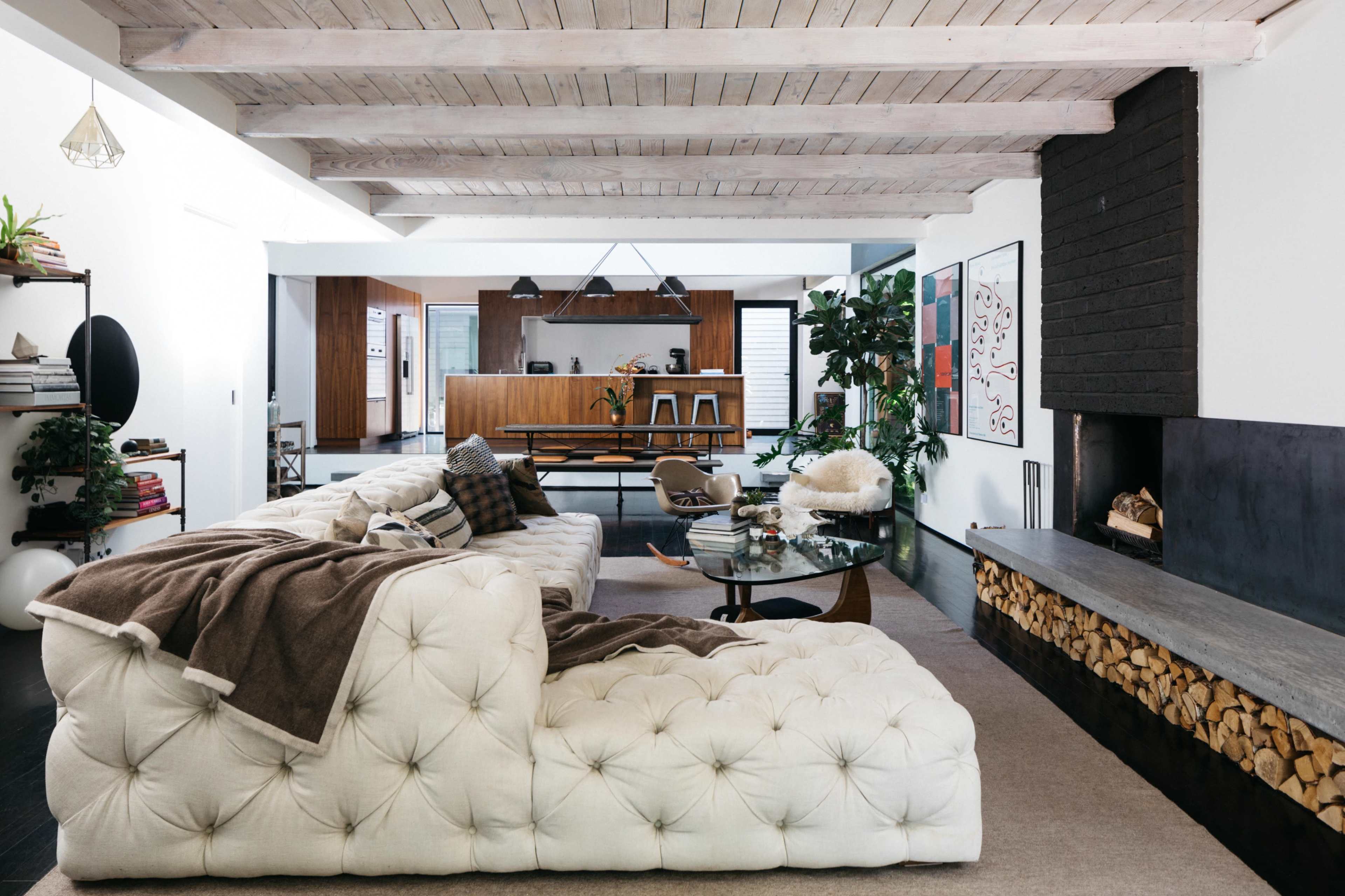 A modern living room features a large tufted white sectional sofa, a round glass coffee table, and a minimalist black fireplace, with exposed wooden beams overhead and a view of an open kitchen in the background.