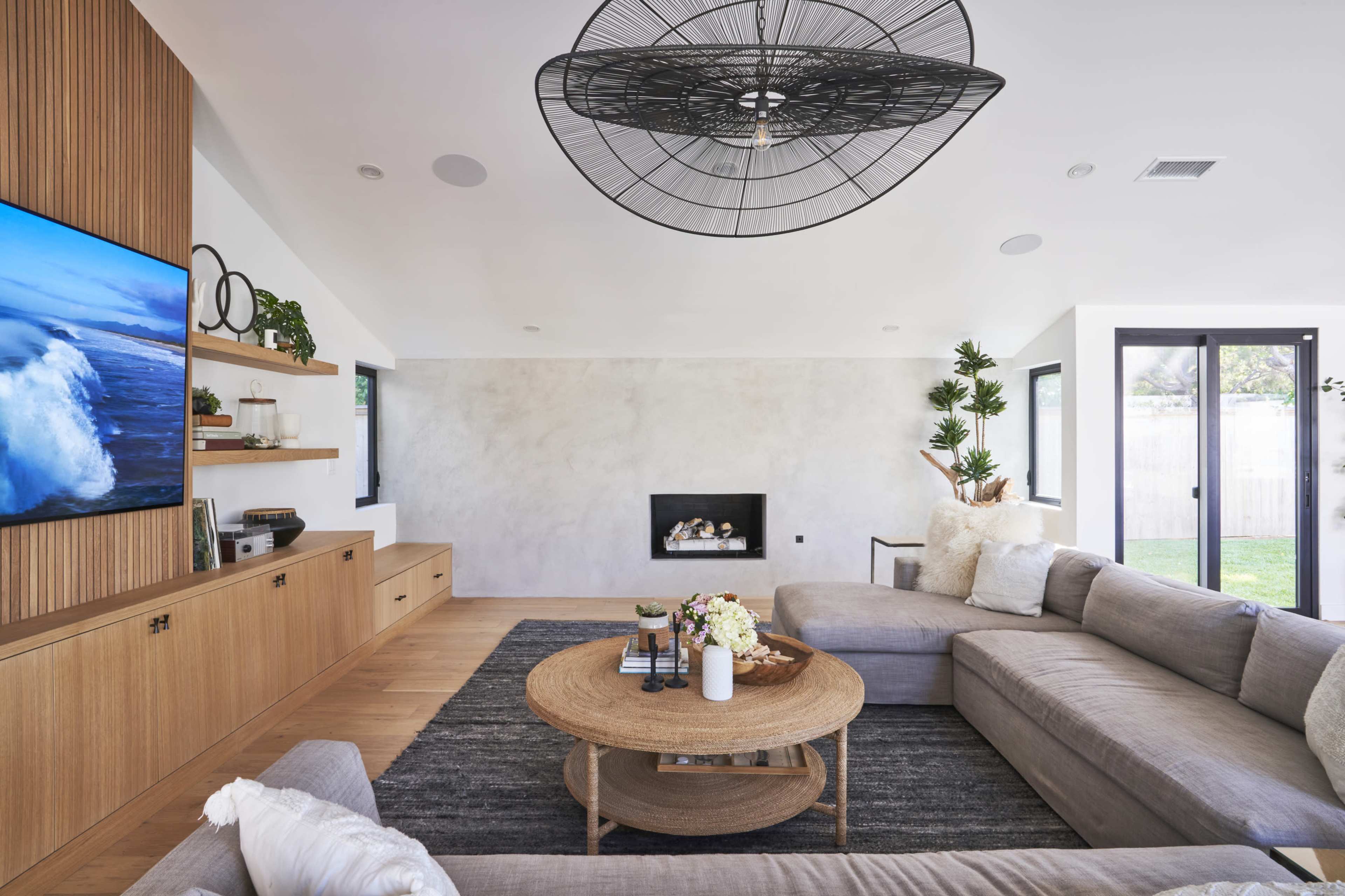 A modern living room with a large sectional sofa, a circular coffee table, a television mounted on a wooden wall unit, and a fireplace between windows.