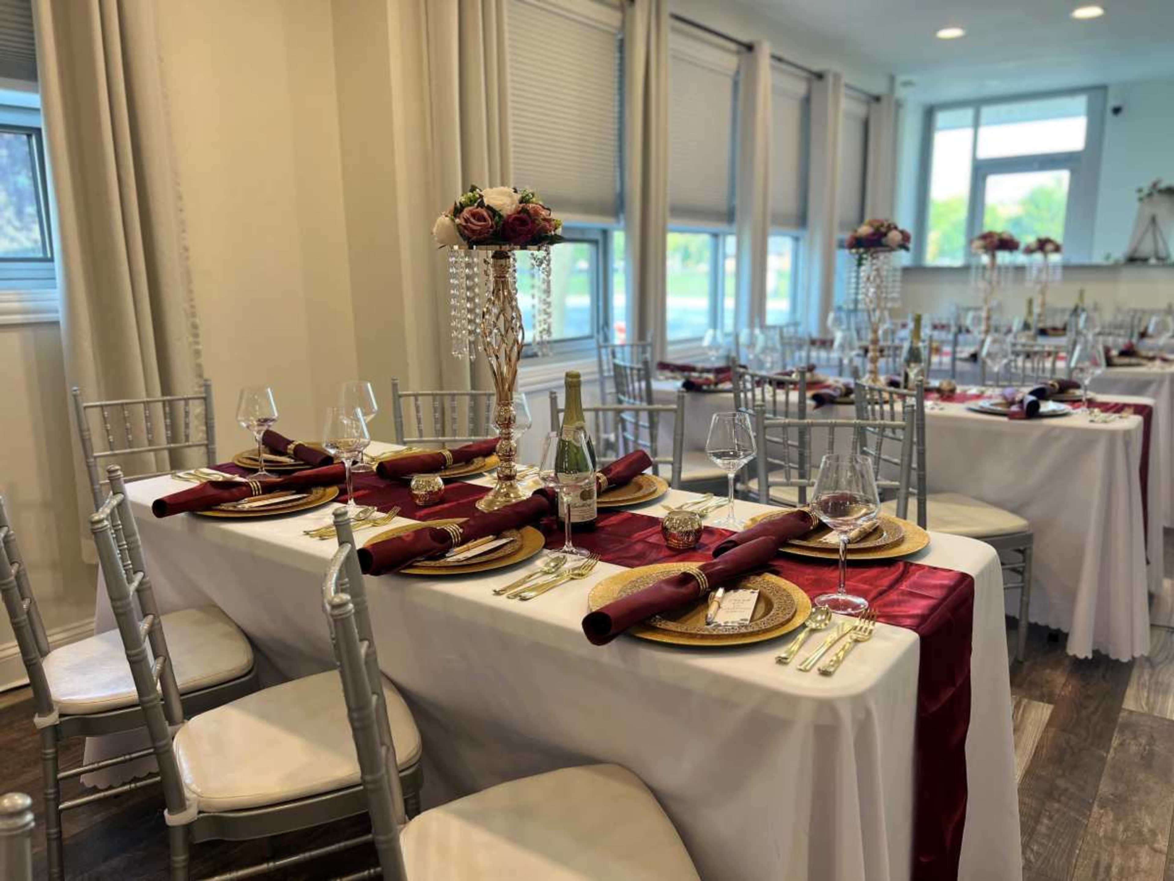 The image shows a banquet hall set for a formal event, featuring elegantly arranged tables with white linens, red accents, fine china, and decorative floral centerpieces.