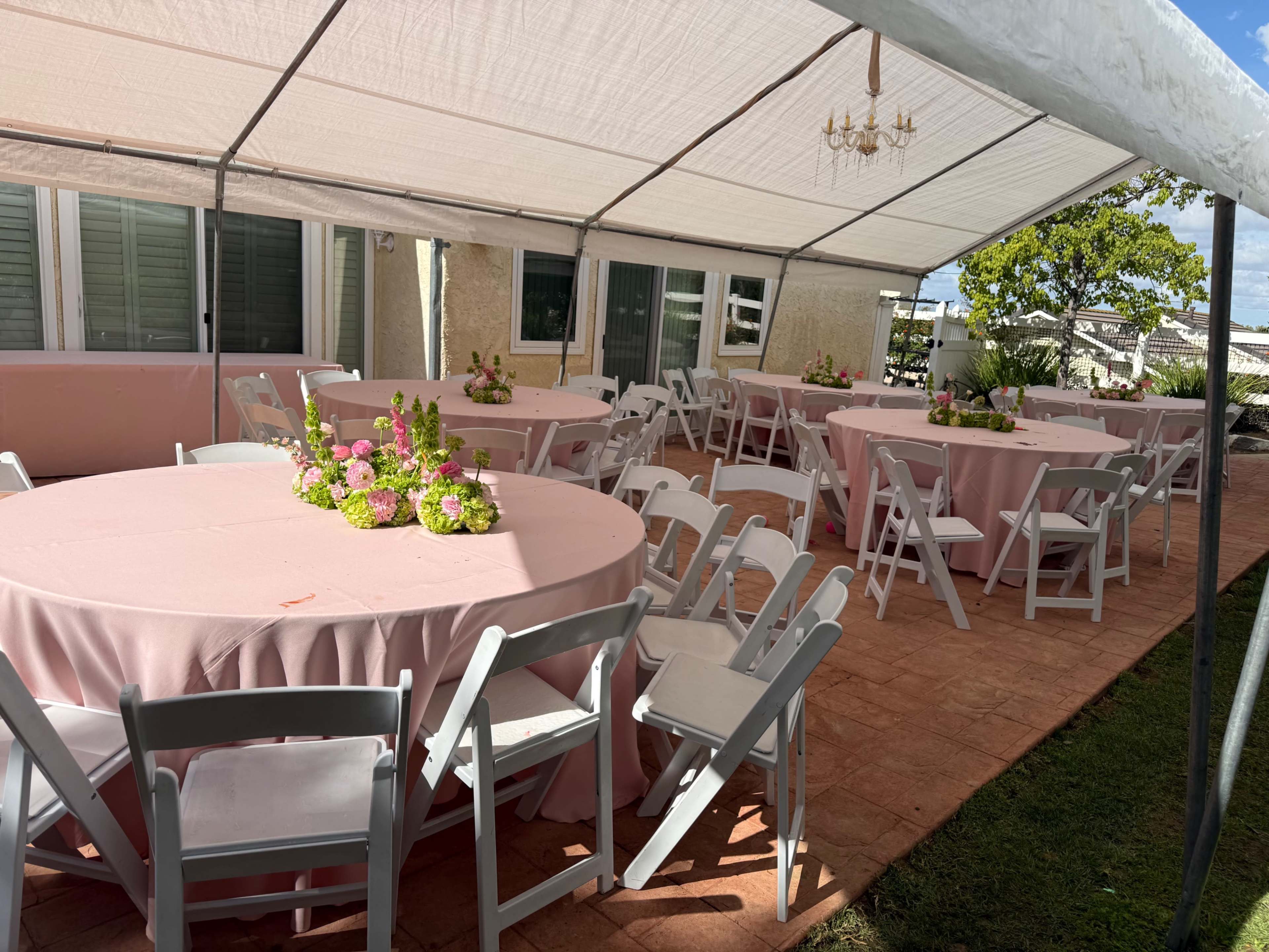 A tented outdoor venue features round tables covered with pink cloth and decorated with floral centerpieces, surrounded by white folding chairs.