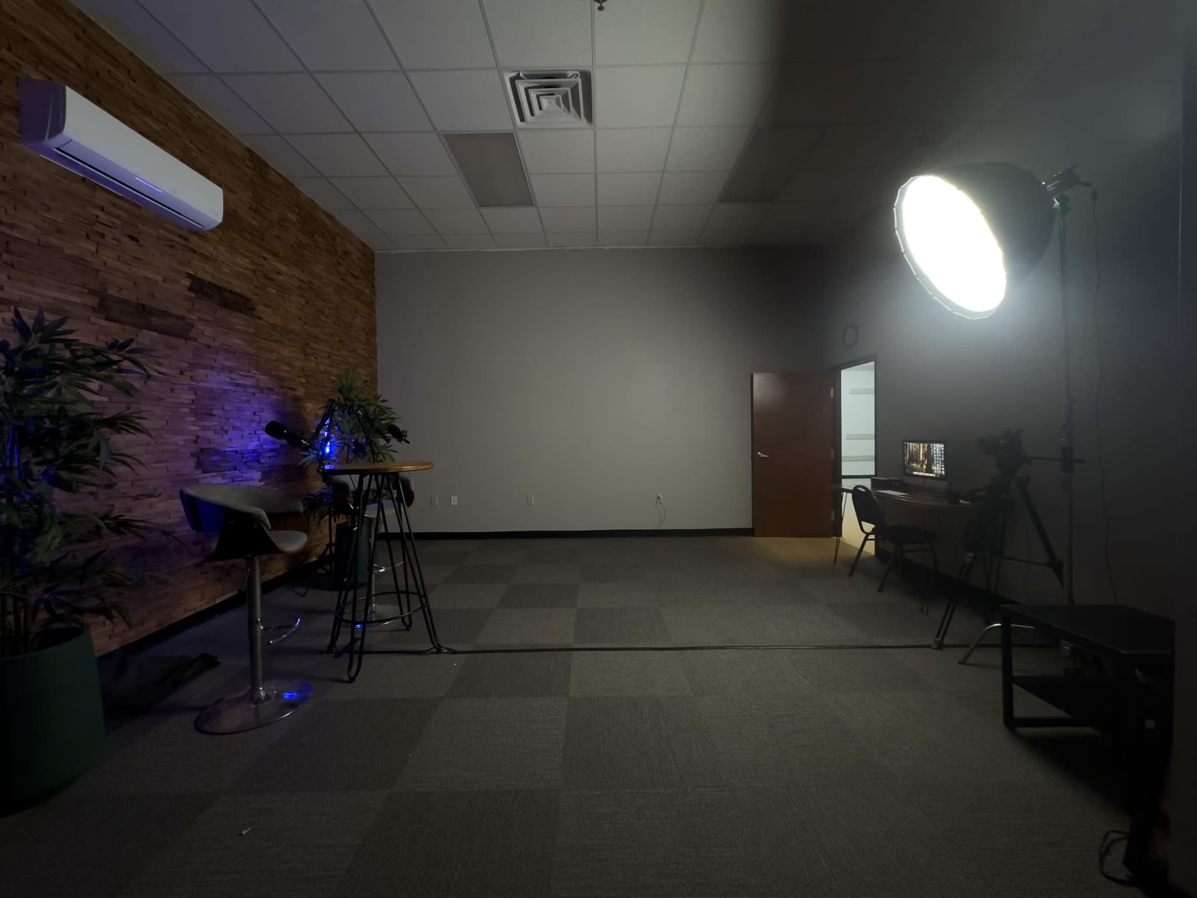 The image shows a dimly lit studio space with a wood-paneled wall, a bar-height table with stools, and a large softbox light in one corner, along with a camera setup and a computer setup on the opposite side.
