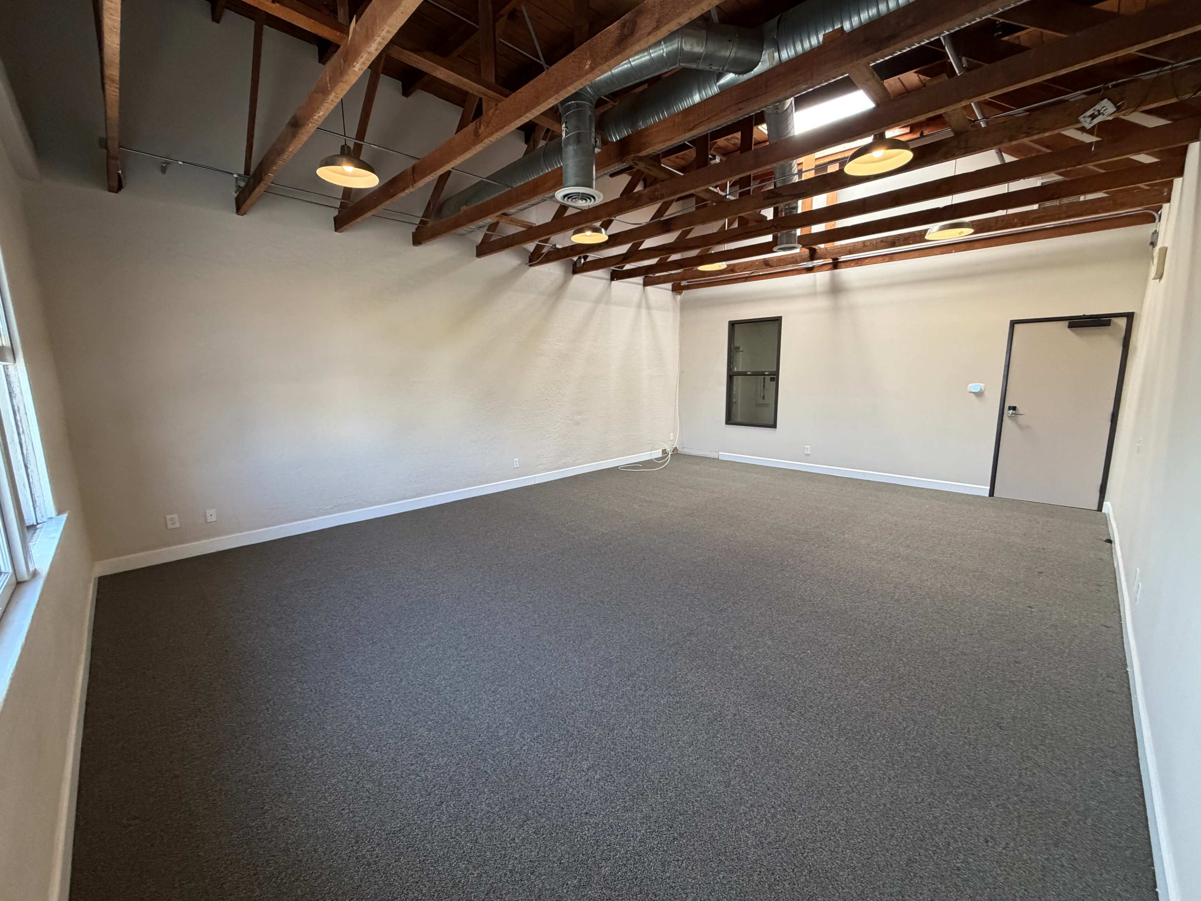 The image shows a spacious room with a high ceiling, exposed wooden beams, and gray carpeting, featuring a single door and window.