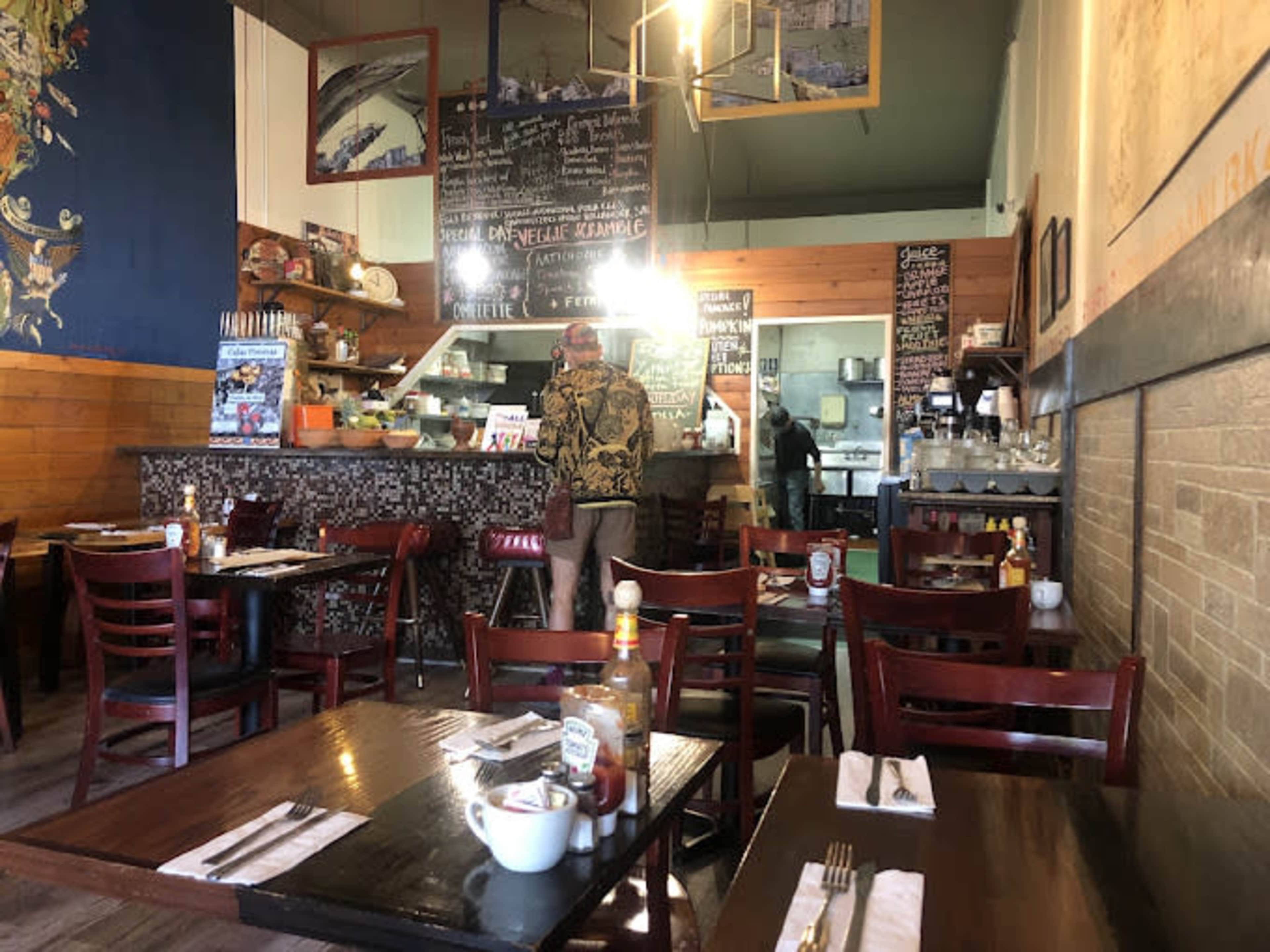 The image shows a cozy restaurant interior with wooden tables and chairs, a counter displaying a menu, and a staff member in the background.