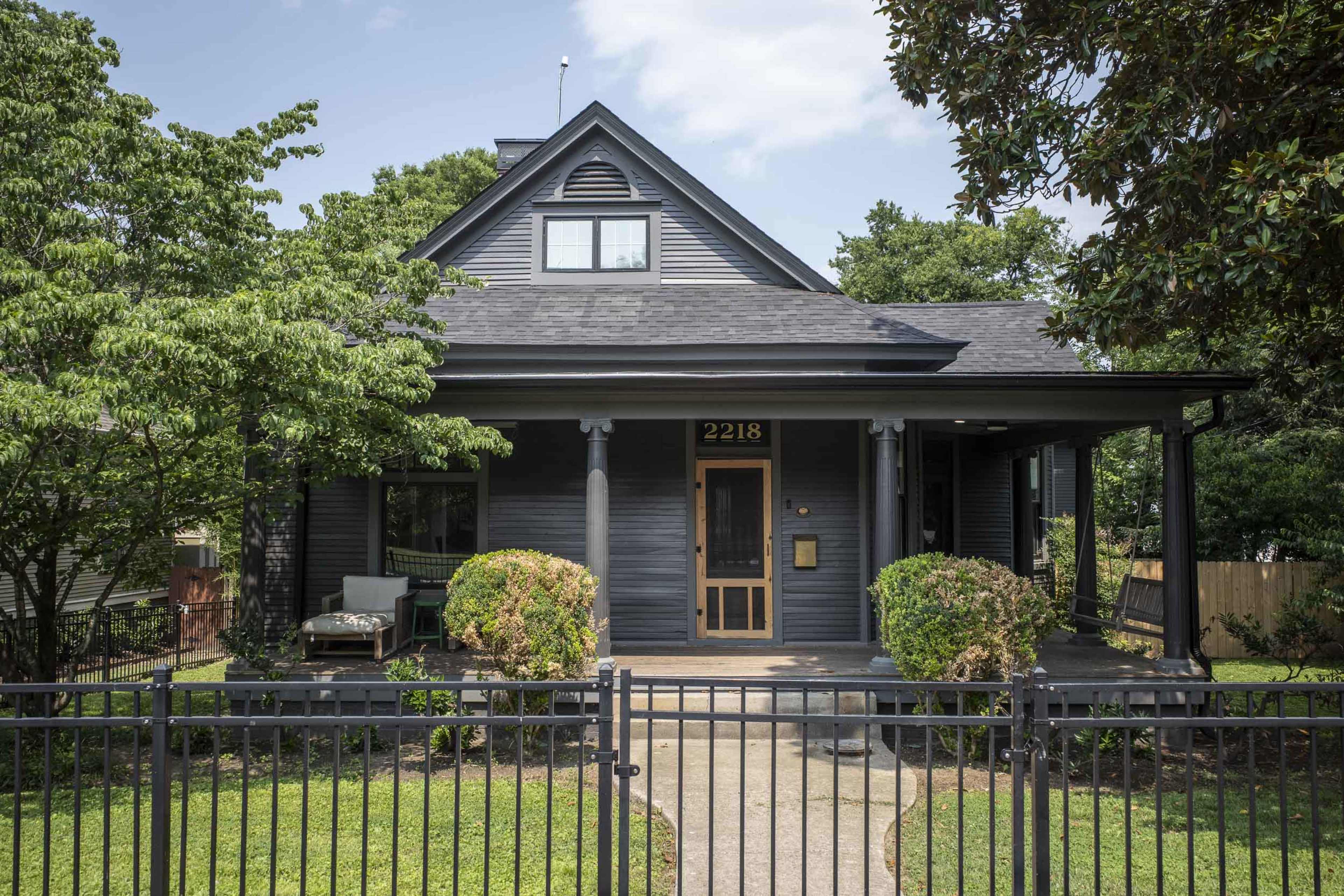 A dark-colored house with a front porch and landscaped greenery is set behind a metal fence.