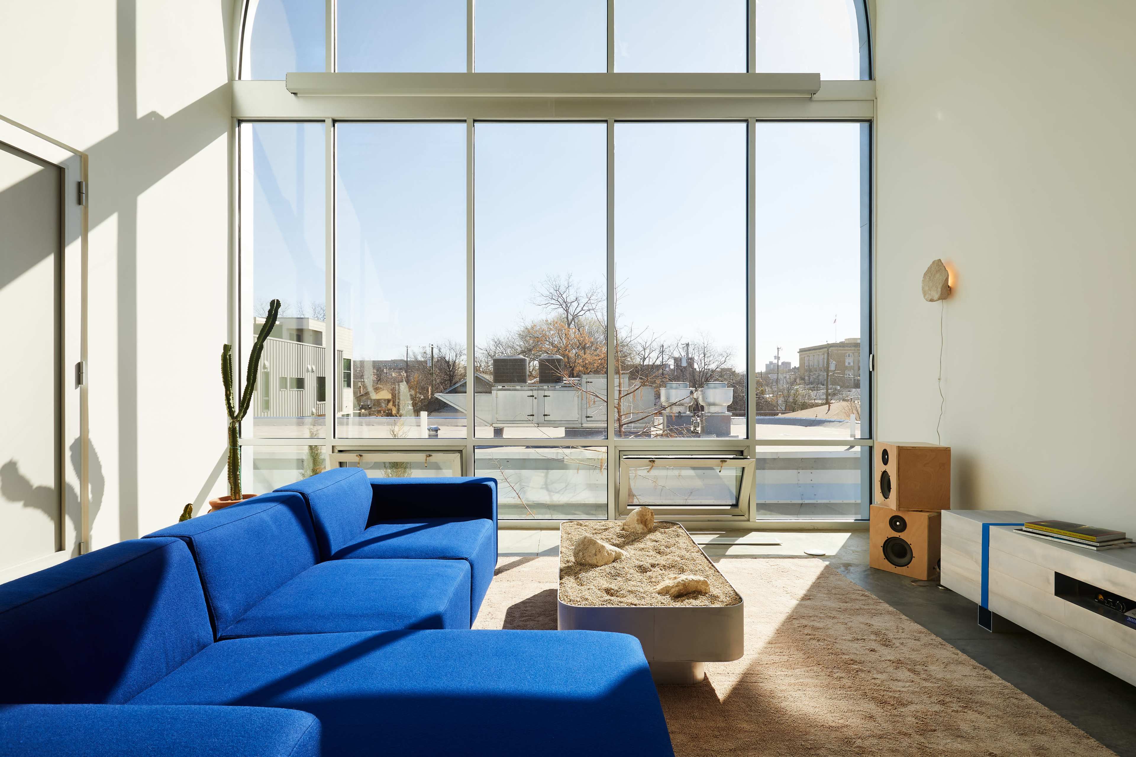 A modern living room features a blue sectional sofa, a low coffee table with decorative stones, and large windows showcasing an urban view.