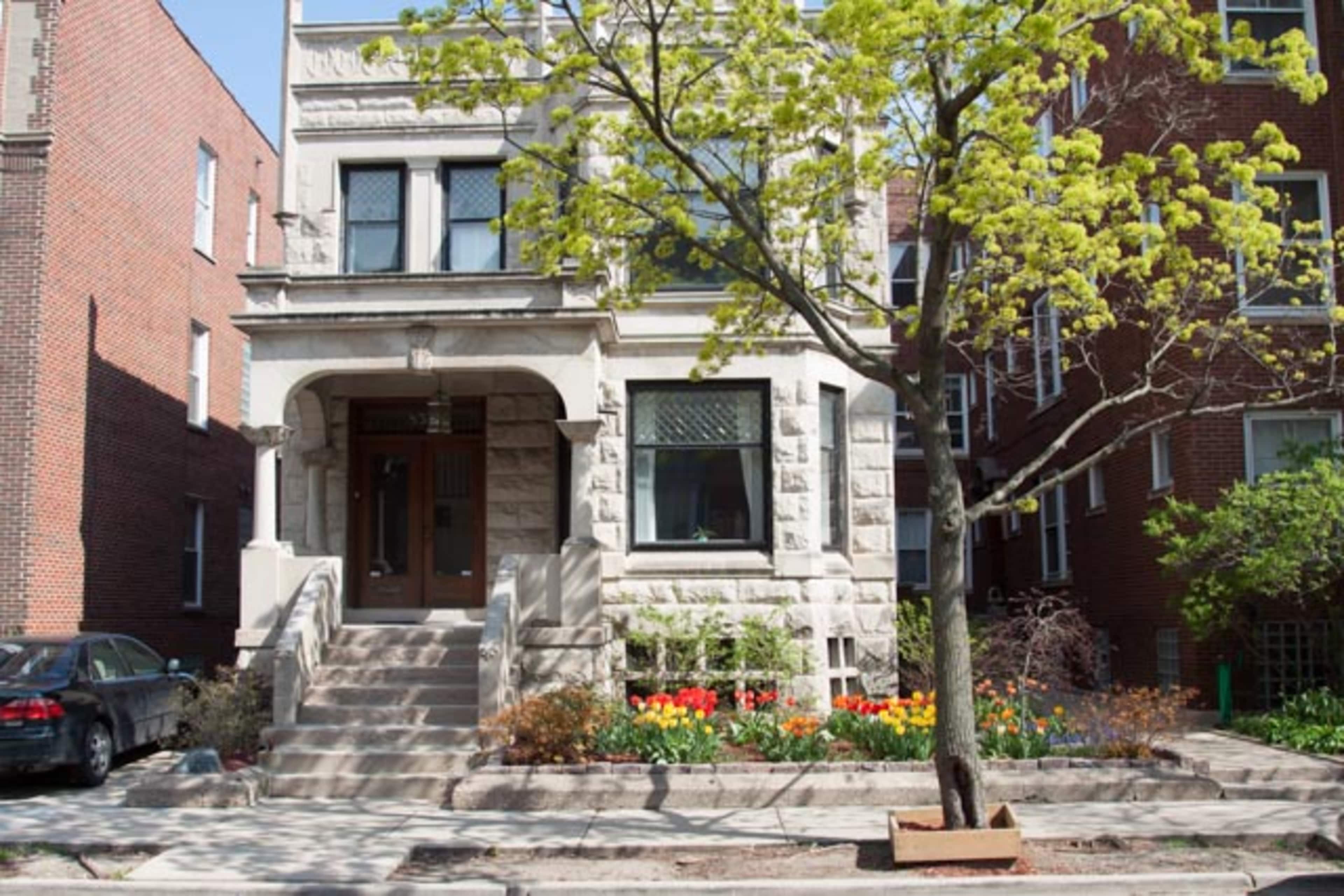 A three-story stone-faced residential building with a front porch is surrounded by flower beds and a small tree.