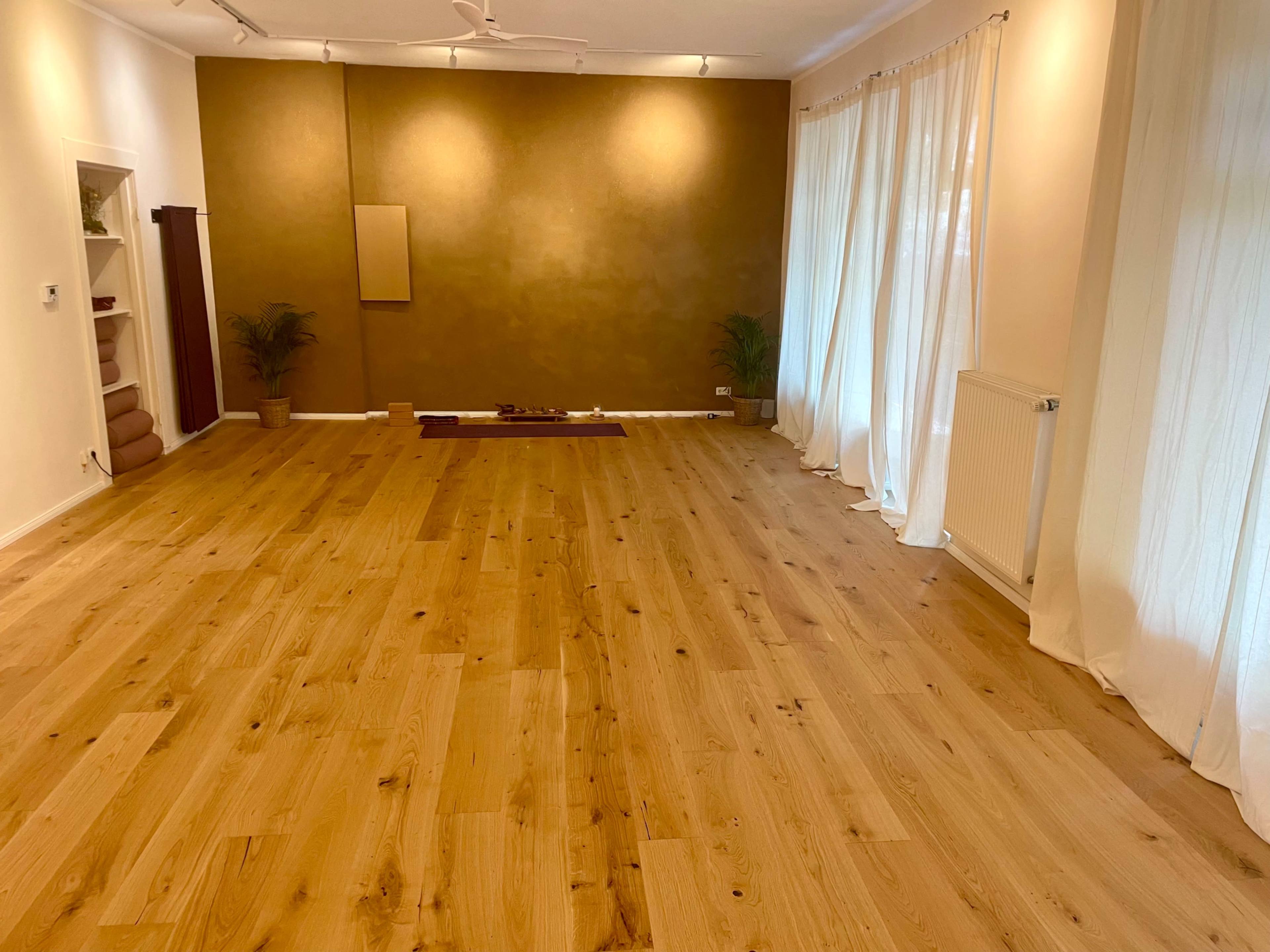 The image shows a spacious room with wooden flooring, a golden accent wall, floor-to-ceiling curtains, and a few decorative plants.