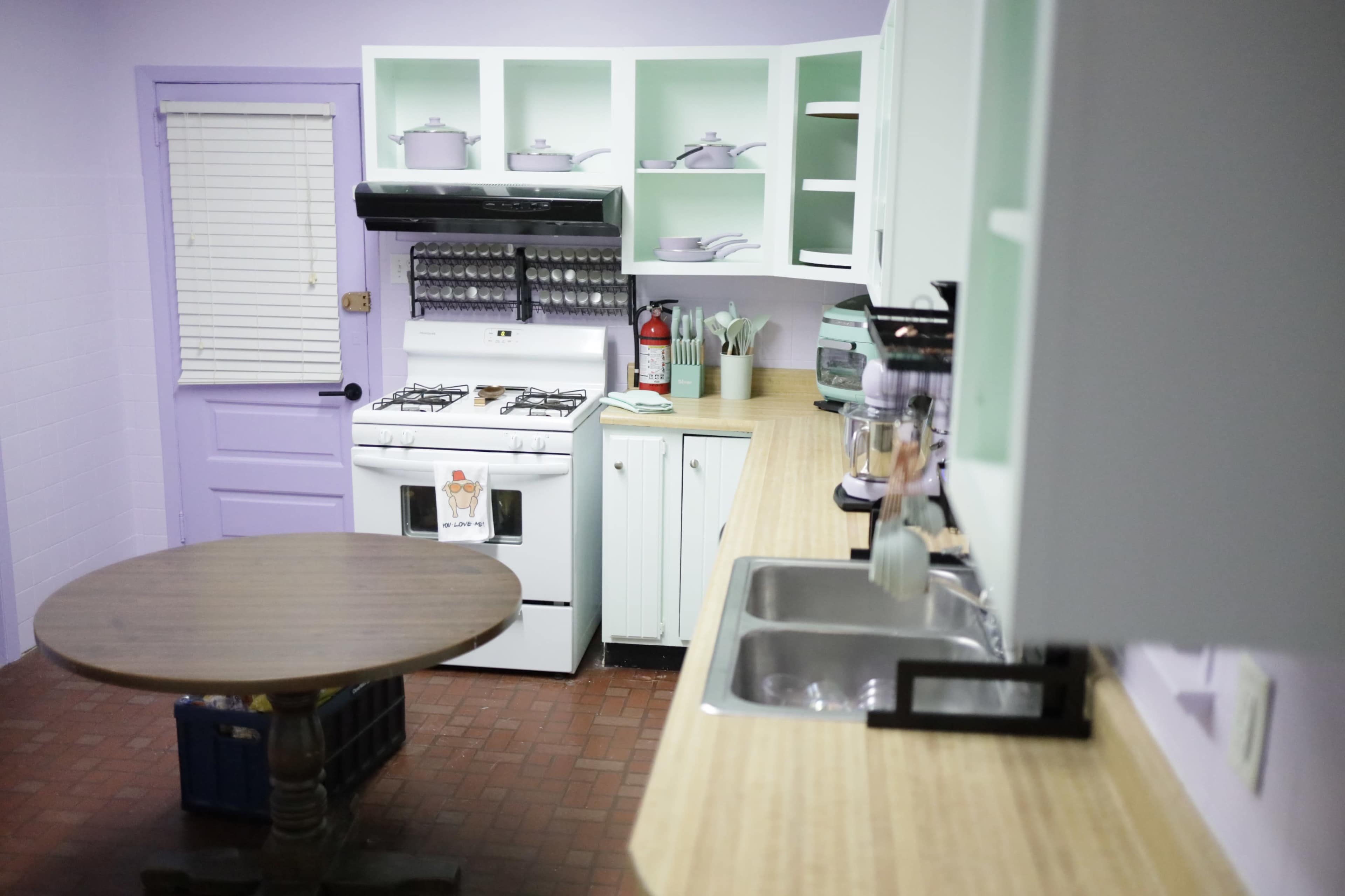 The kitchen features a teal and white color scheme with a wooden table, an oven, and various kitchen utensils on the countertops.