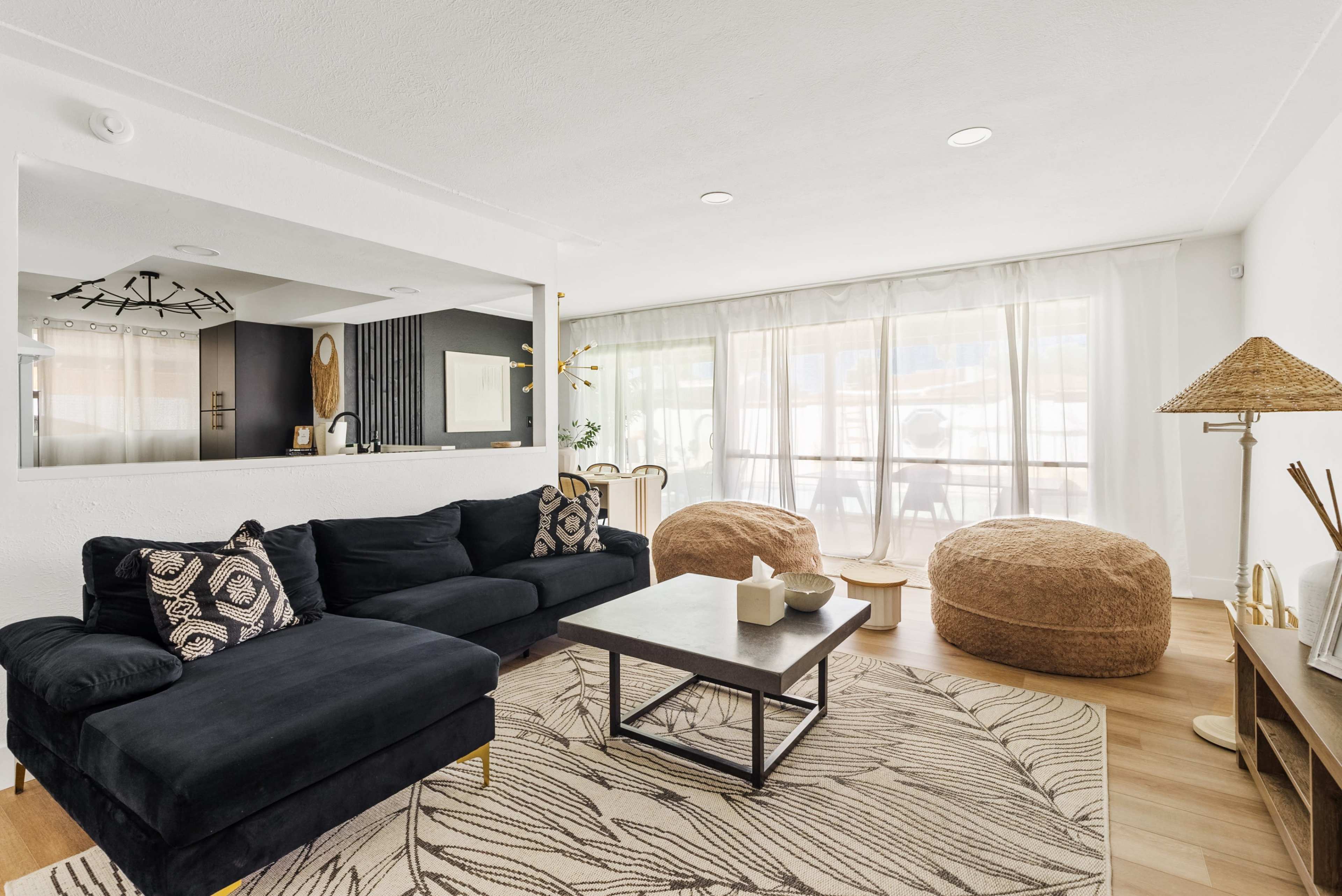 The image shows a modern living room featuring a sectional sofa, a coffee table, and decorative elements against a backdrop of large windows with sheer curtains.