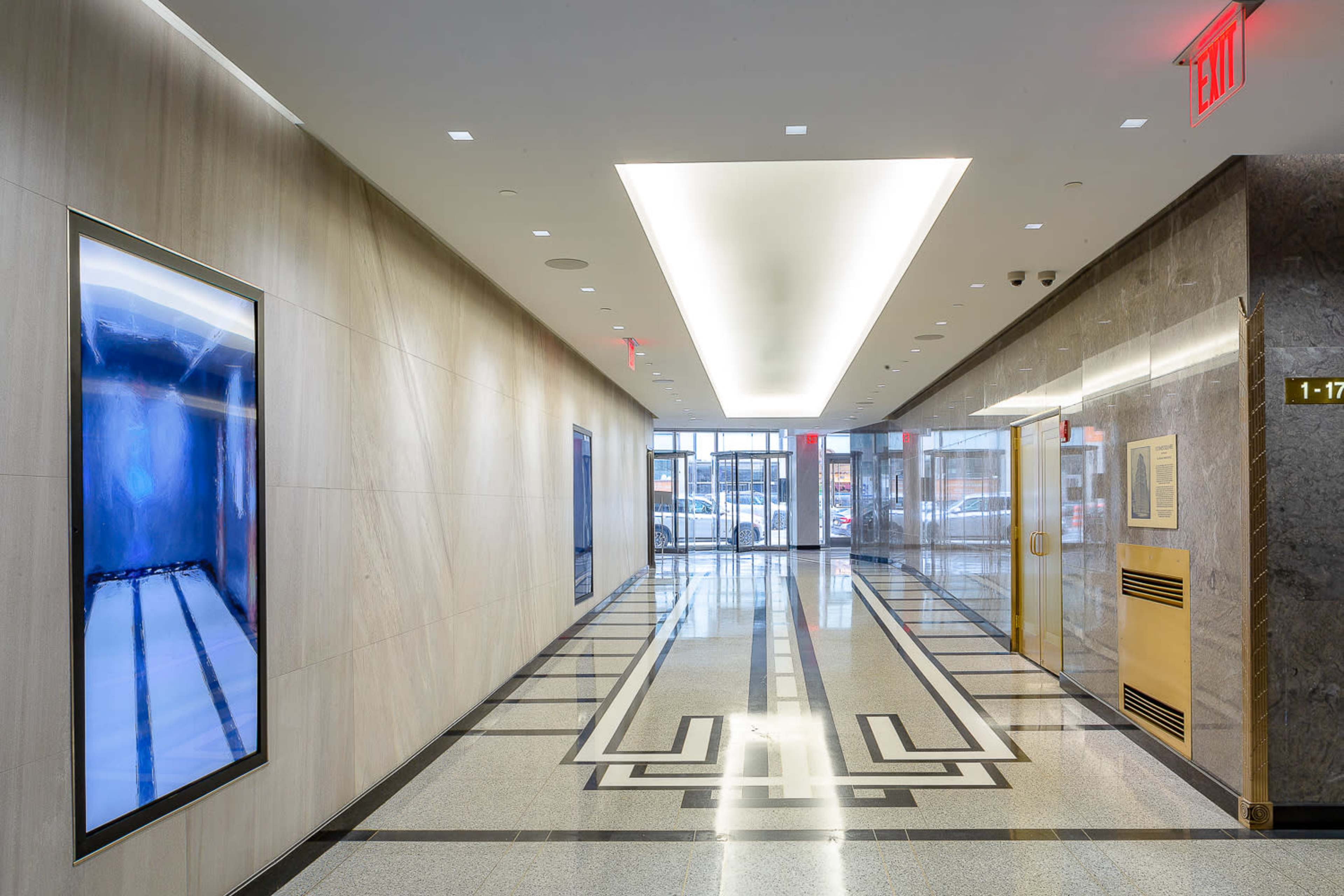 A well-lit hall features large artwork on the walls and sleek tiled flooring, leading to an entrance with glass doors.