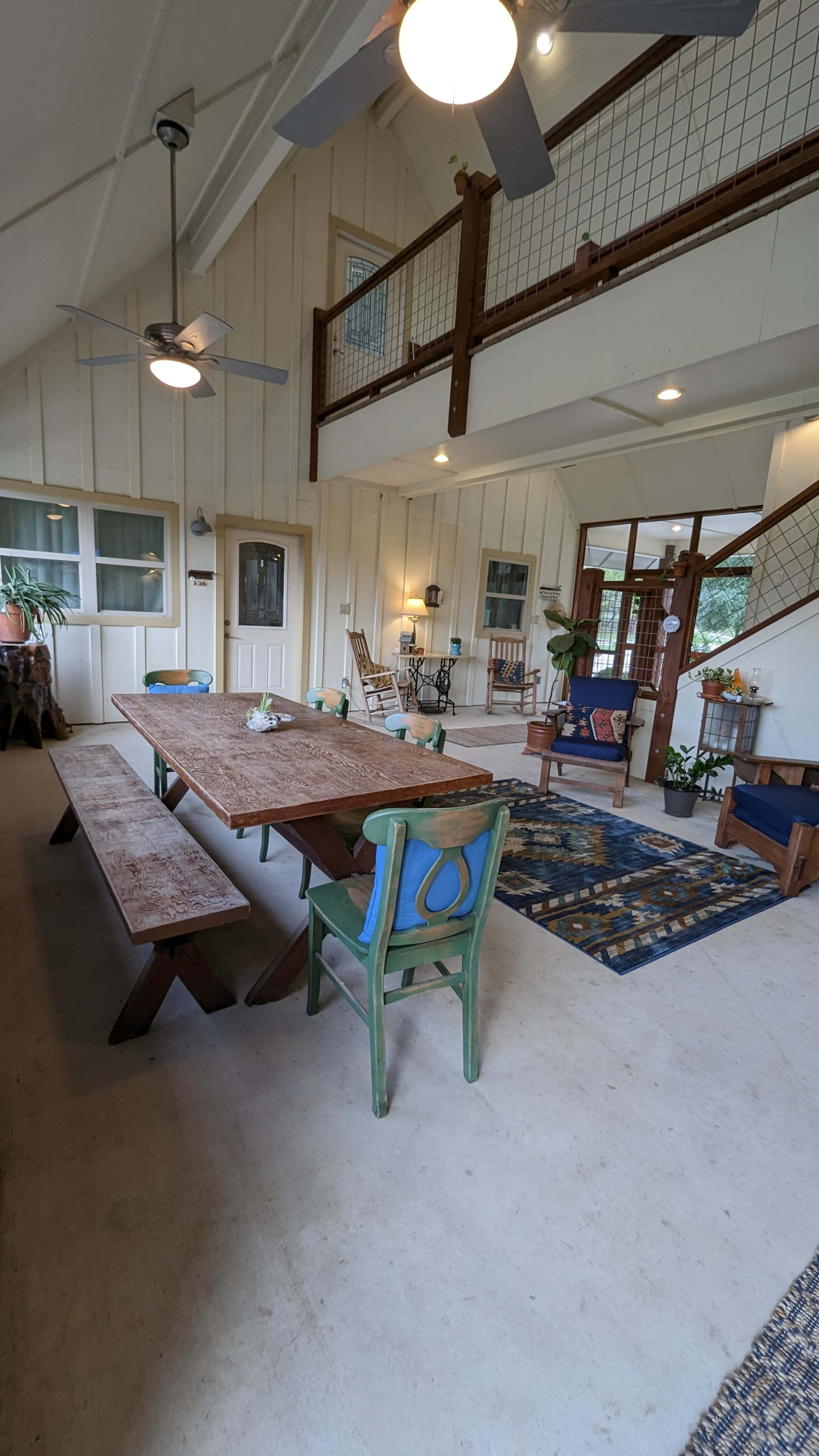 A spacious, well-lit interior with a large wooden dining table surrounded by benches, colorful chairs, and various decorative elements, including plants and rugs, in an open-plan layout.