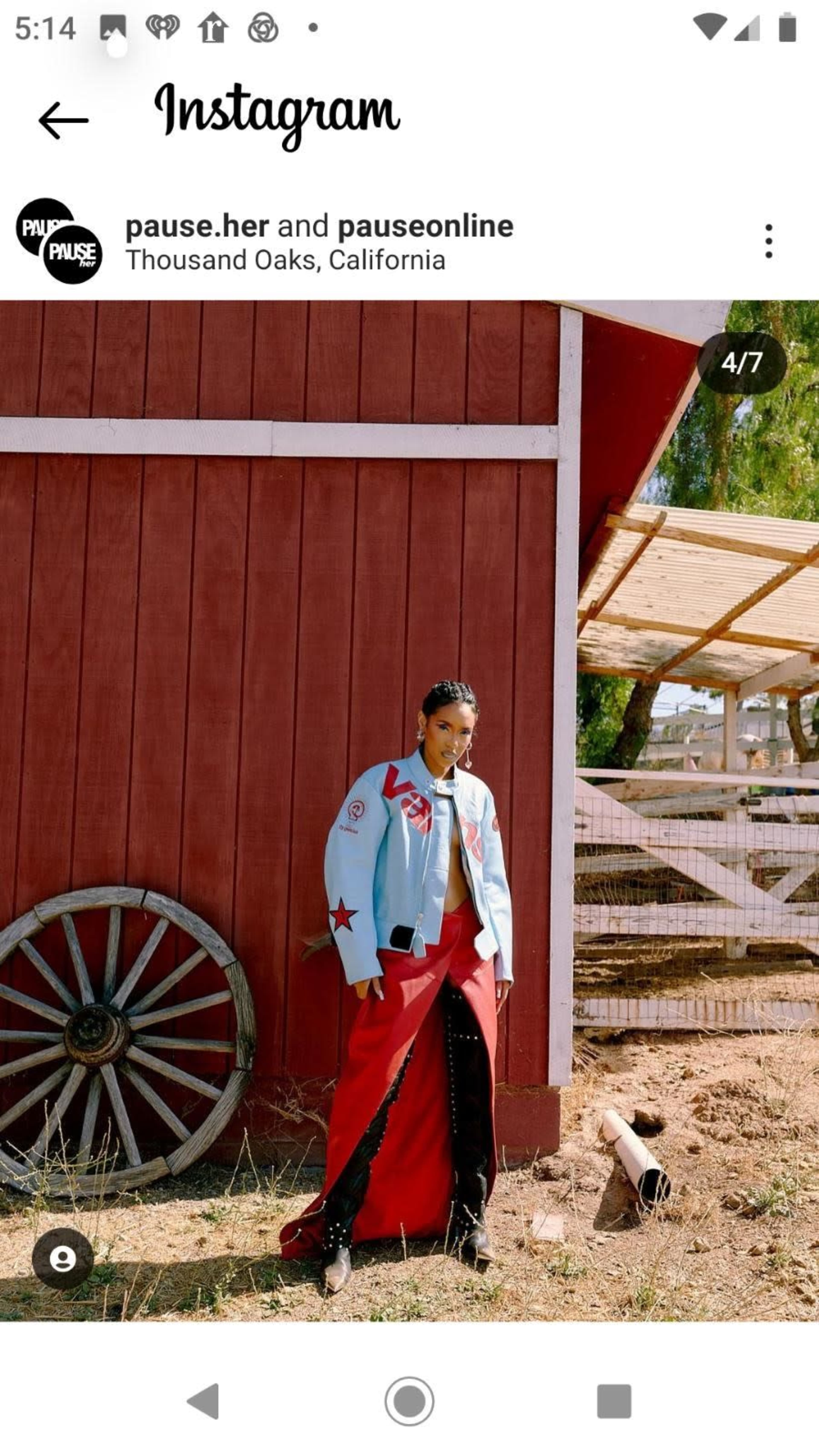 A person stands beside a red barn, wearing a blue jacket with stars and a long red skirt.