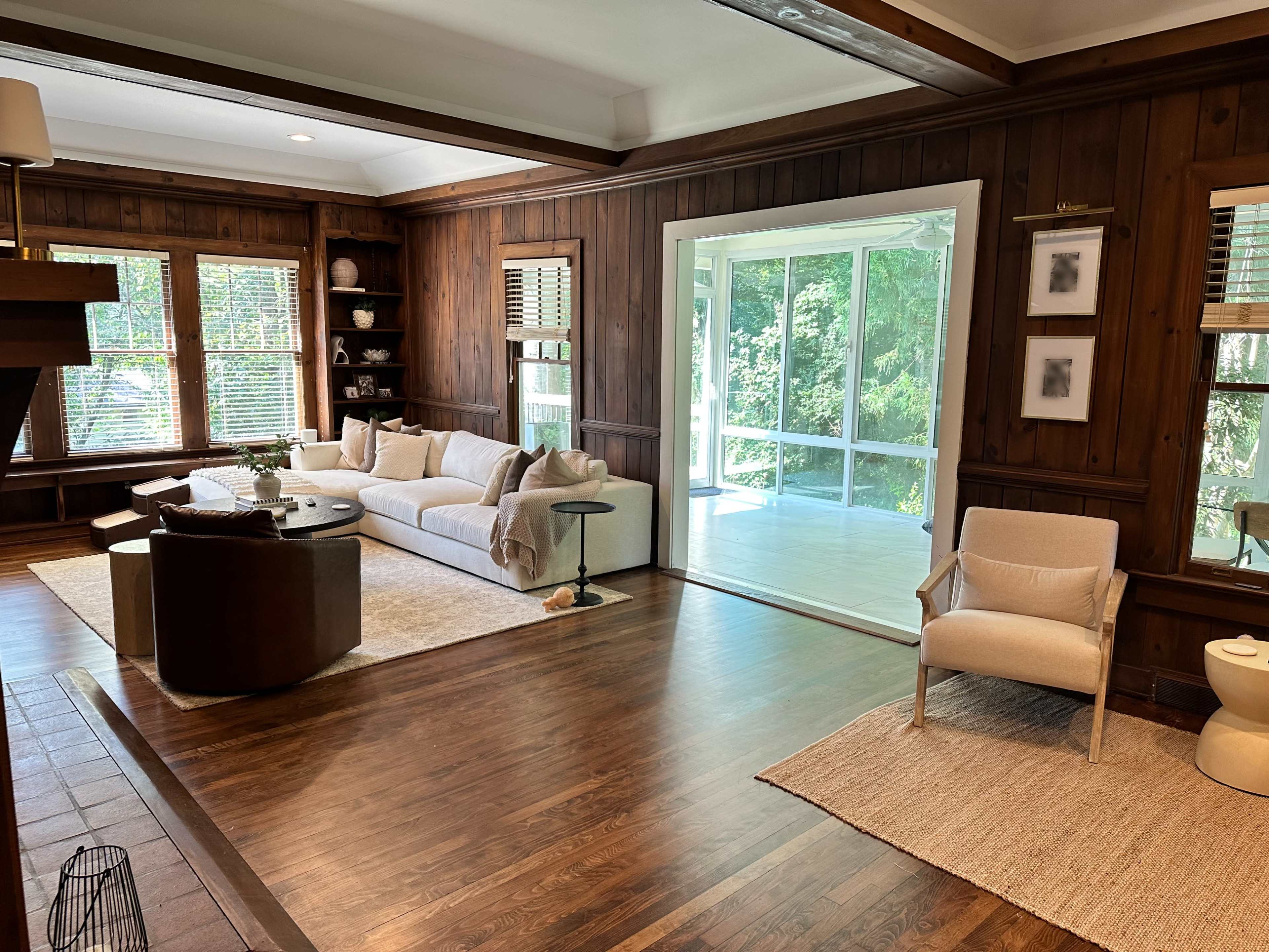 The image shows a spacious living room with wooden paneling, a large sectional sofa, a coffee table, and a view of a sunlit porch through large glass doors.
