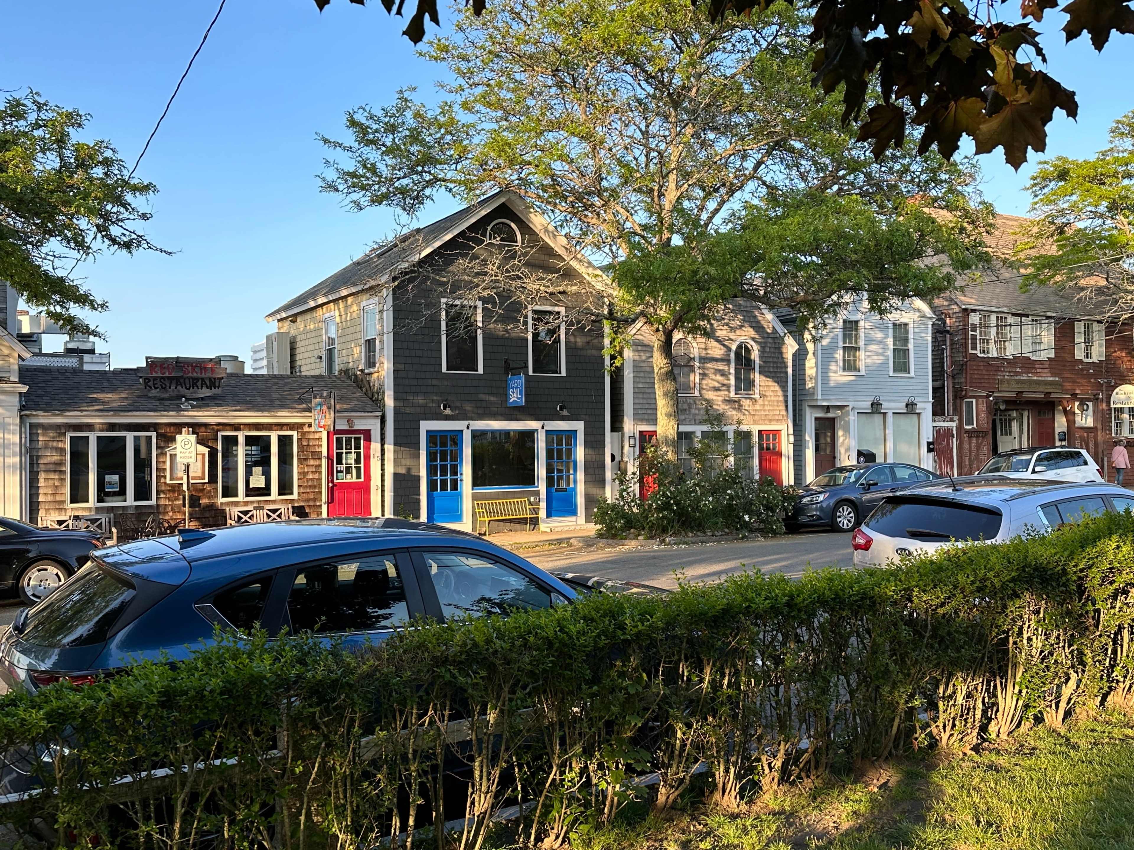 A row of colorful buildings with storefronts and residential units, lined along a street next to parked cars and a hedge.