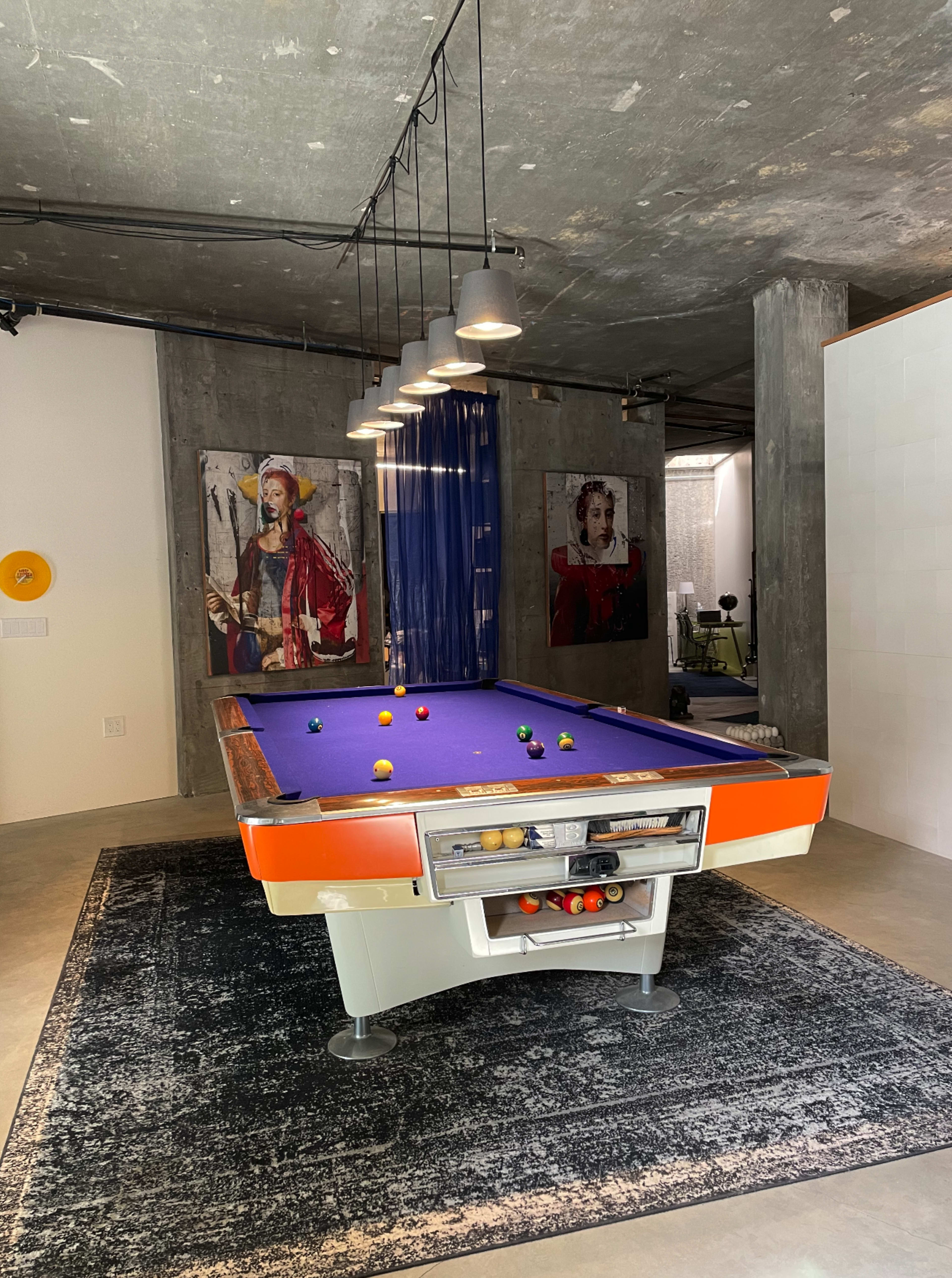 The image shows a modern interior featuring a purple pool table surrounded by a patterned rug, with two large framed artworks displayed on the concrete walls above the table.