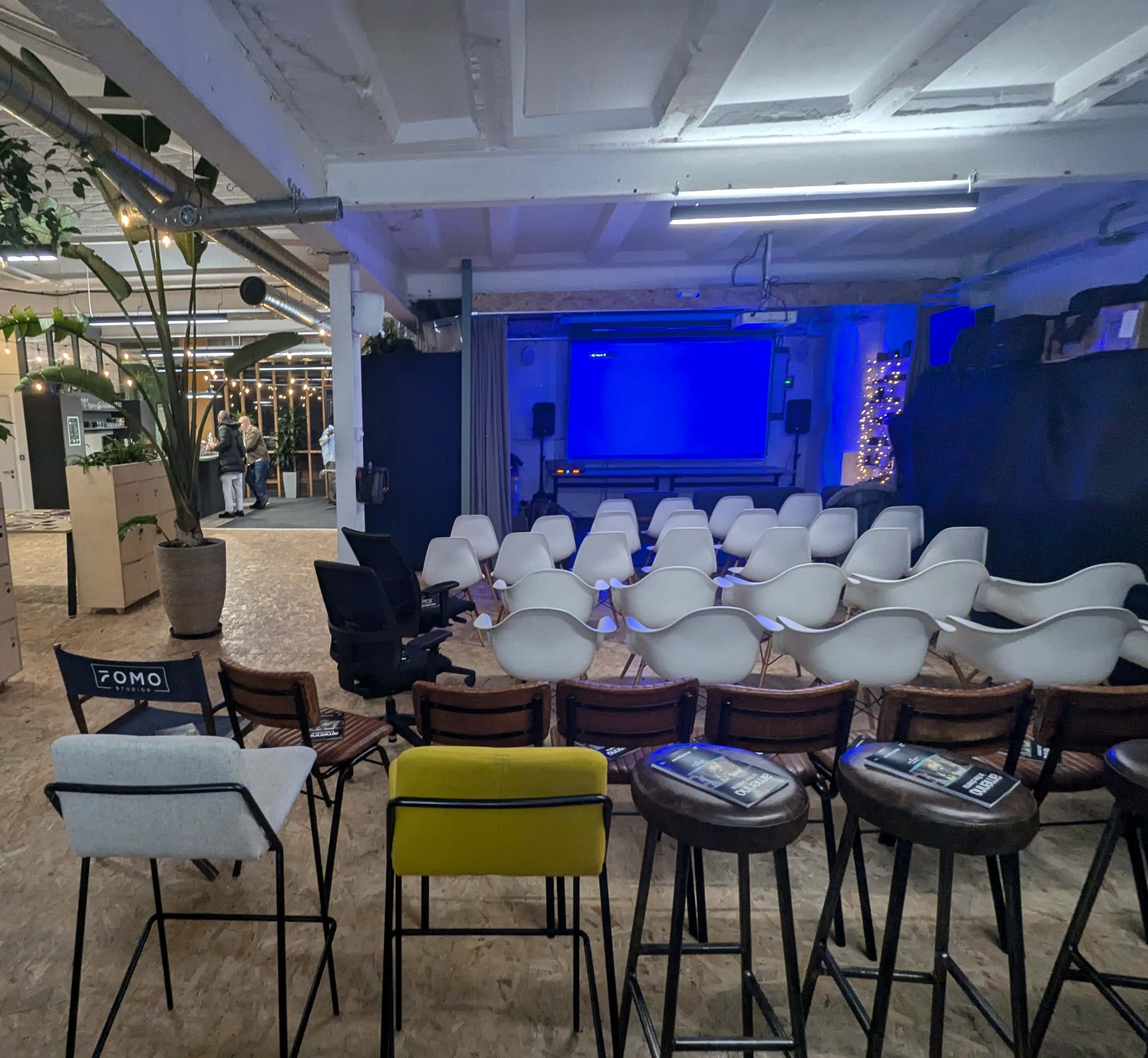 The image shows a spacious indoor venue arranged with rows of white chairs facing a blank blue screen, with some plants and a few tables in the background.