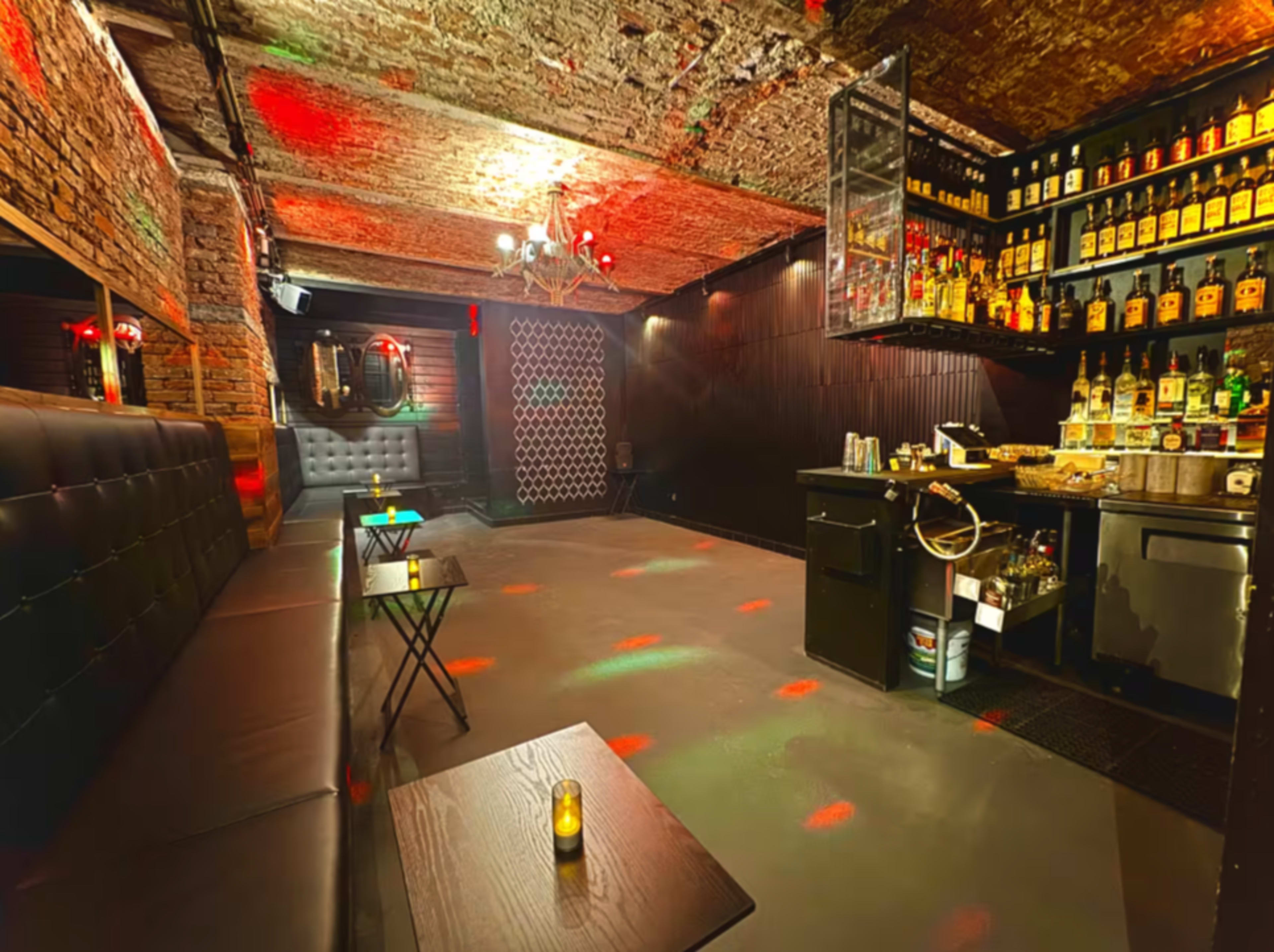 The image shows a dimly lit bar with stone walls, a well-stocked drink shelf, and black seating arranged around small tables.