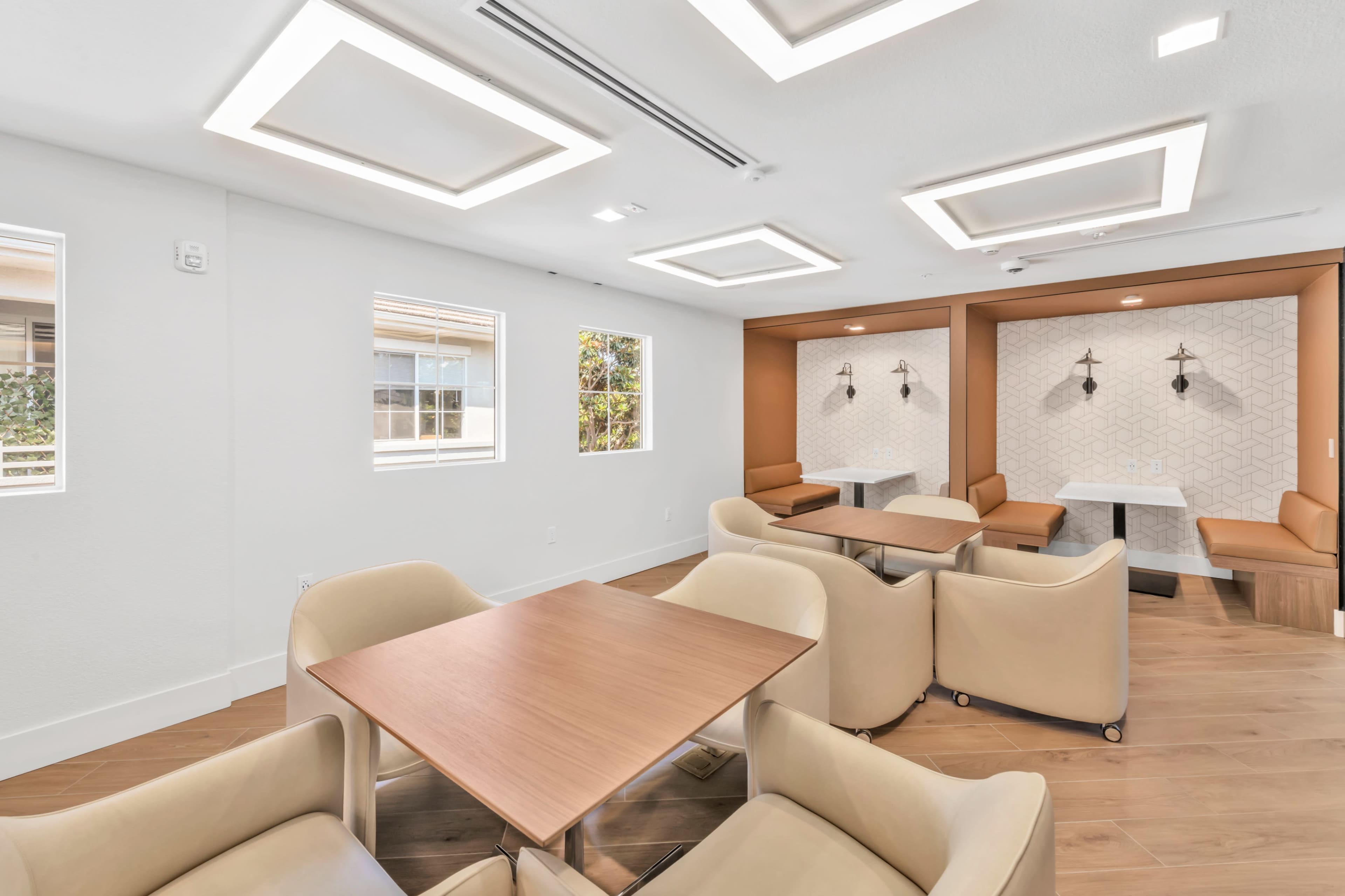 The image shows a modern lounge area with light wooden flooring, four cream-colored armchairs around a wooden table, and two cozy seating arrangements against an orange accent wall.