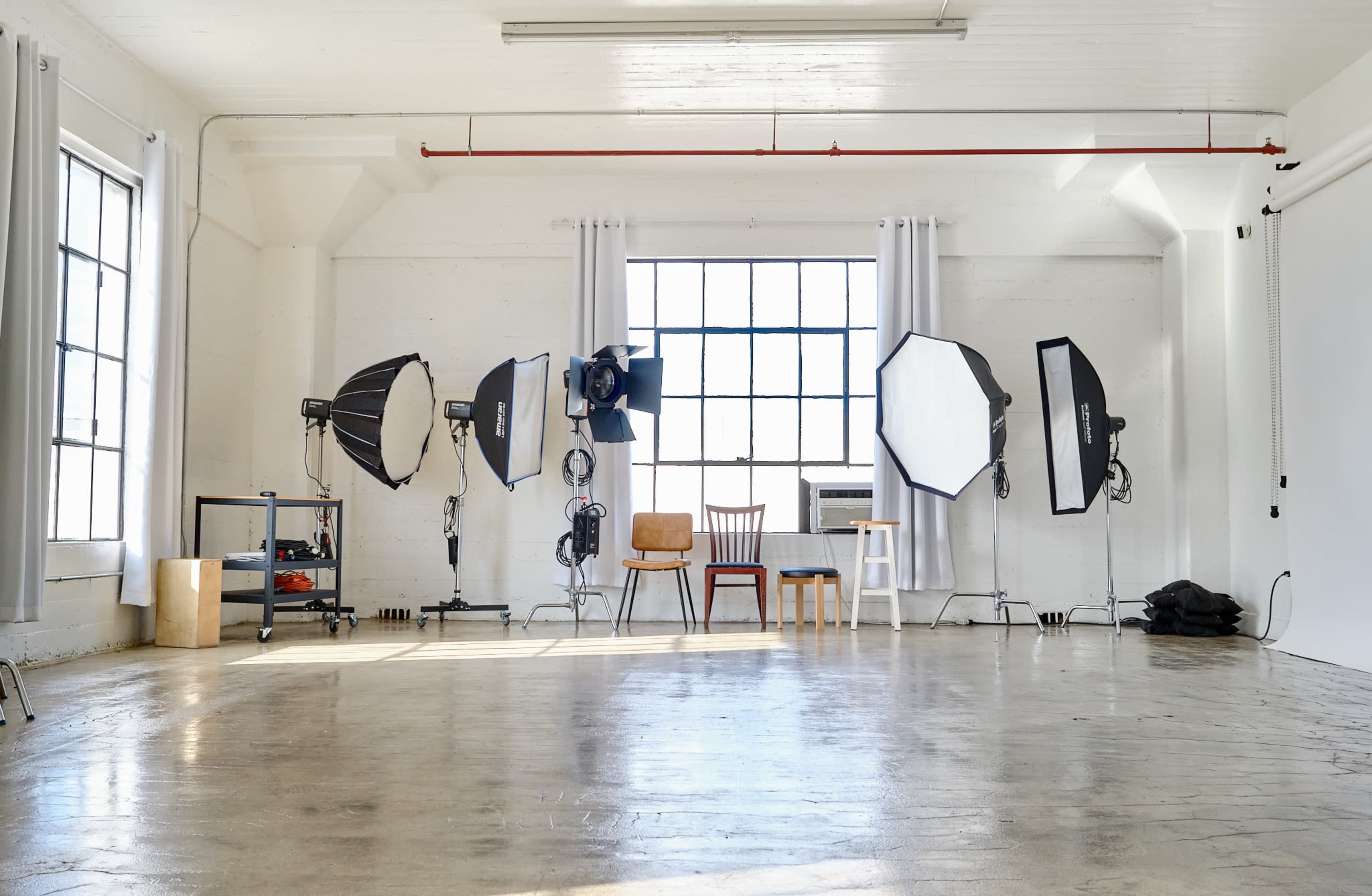 A bright photography studio features several light stands and softboxes arranged in front of large windows, with three chairs positioned in the center.