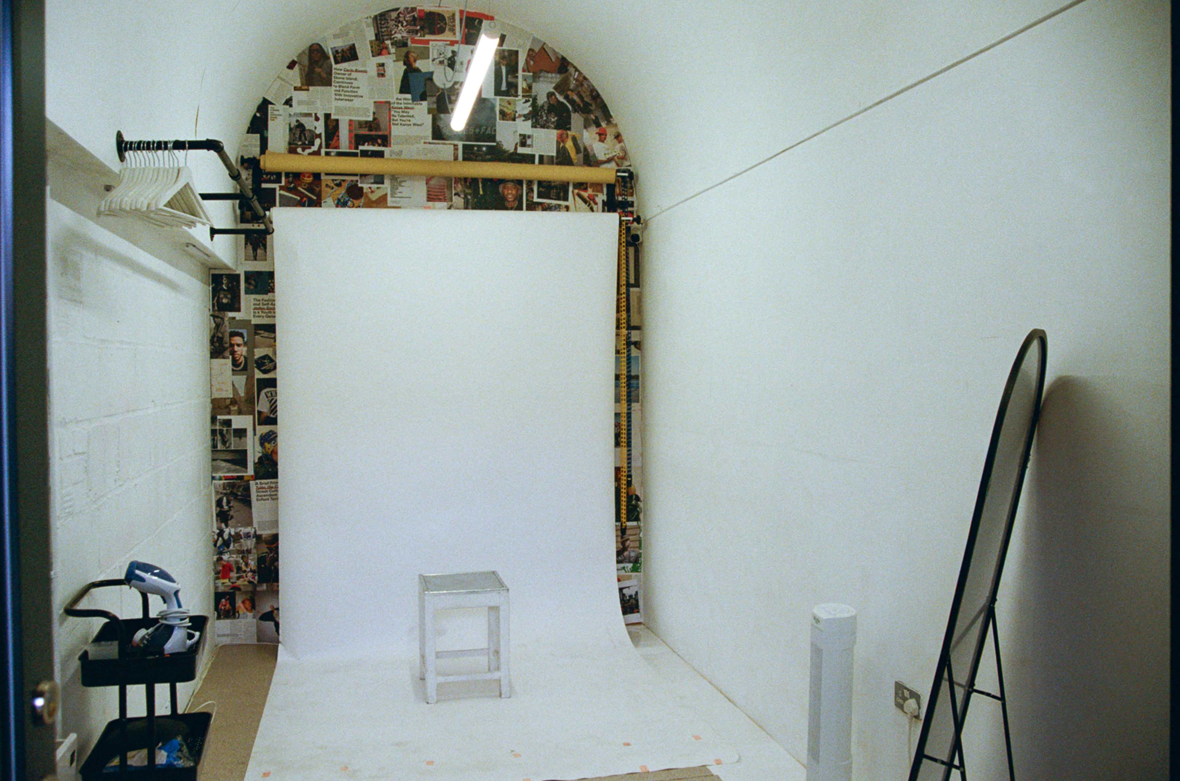 The image shows a small, minimalist photo studio with a white backdrop, a stool, and collaged walls.
