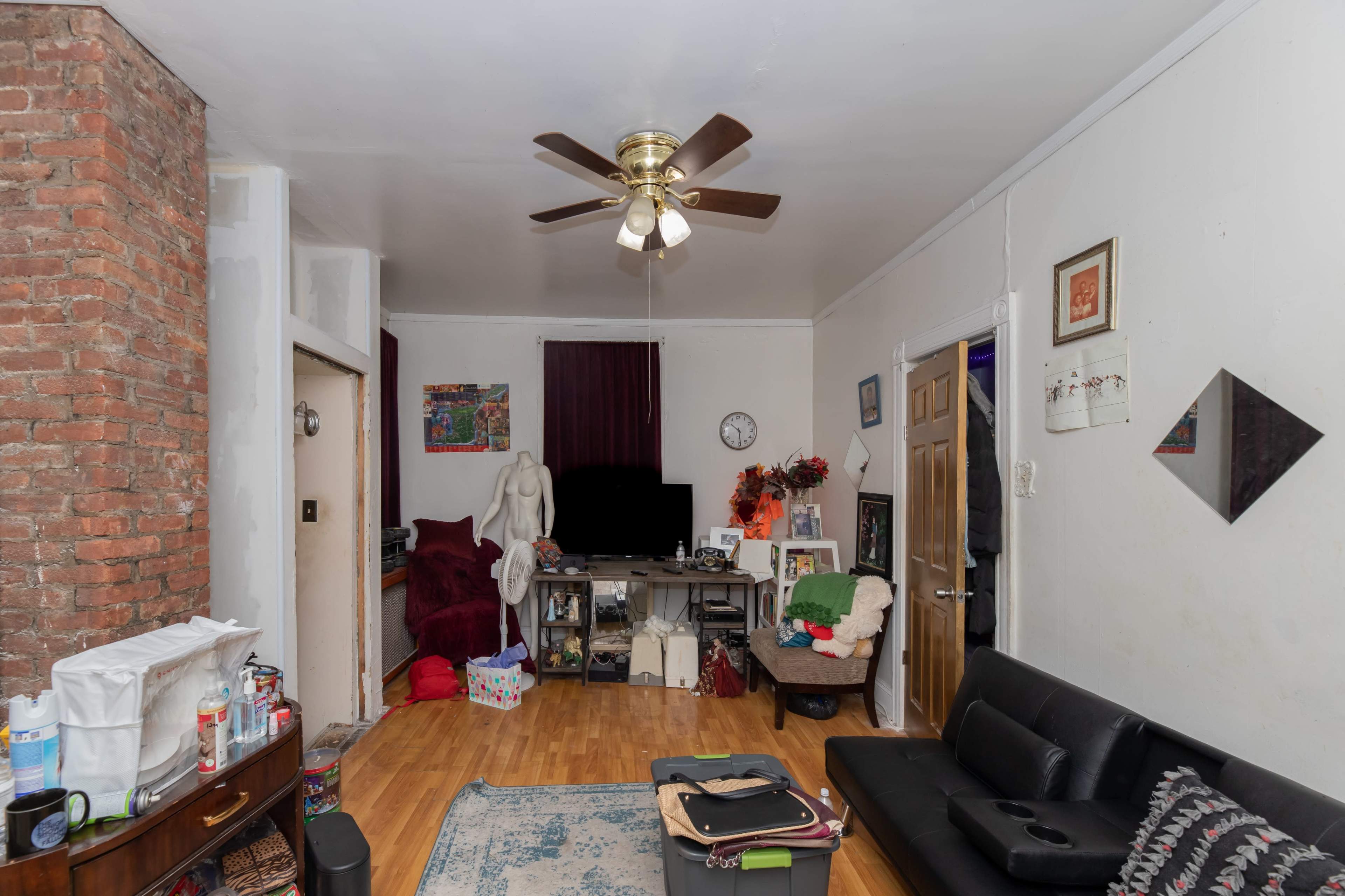 The image shows a living room with a brick wall, a ceiling fan, a black sofa, and a television setup surrounded by various items.