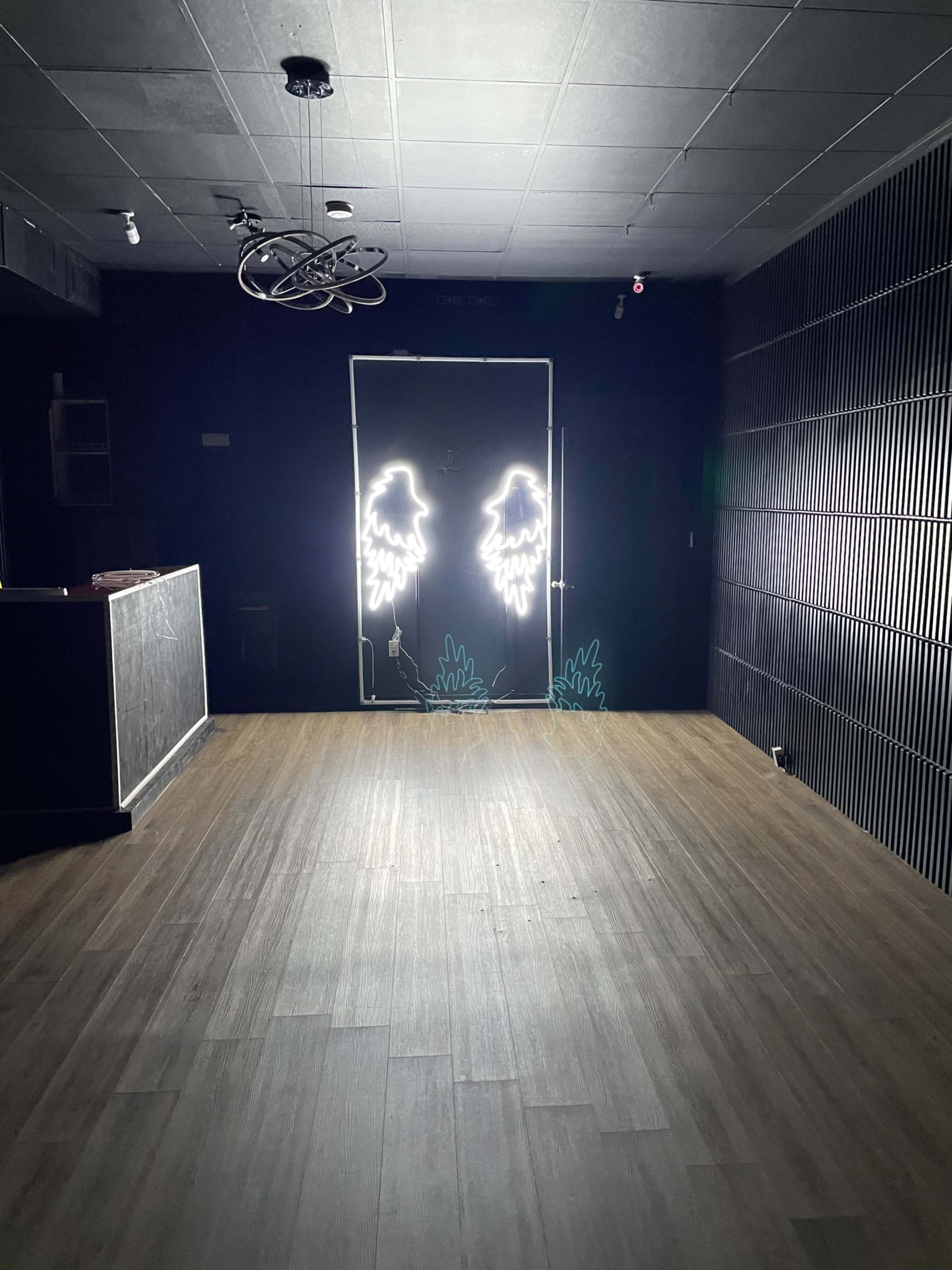 The image shows an empty room with a dark interior featuring a door illuminated by neon light shaped like angel wings.