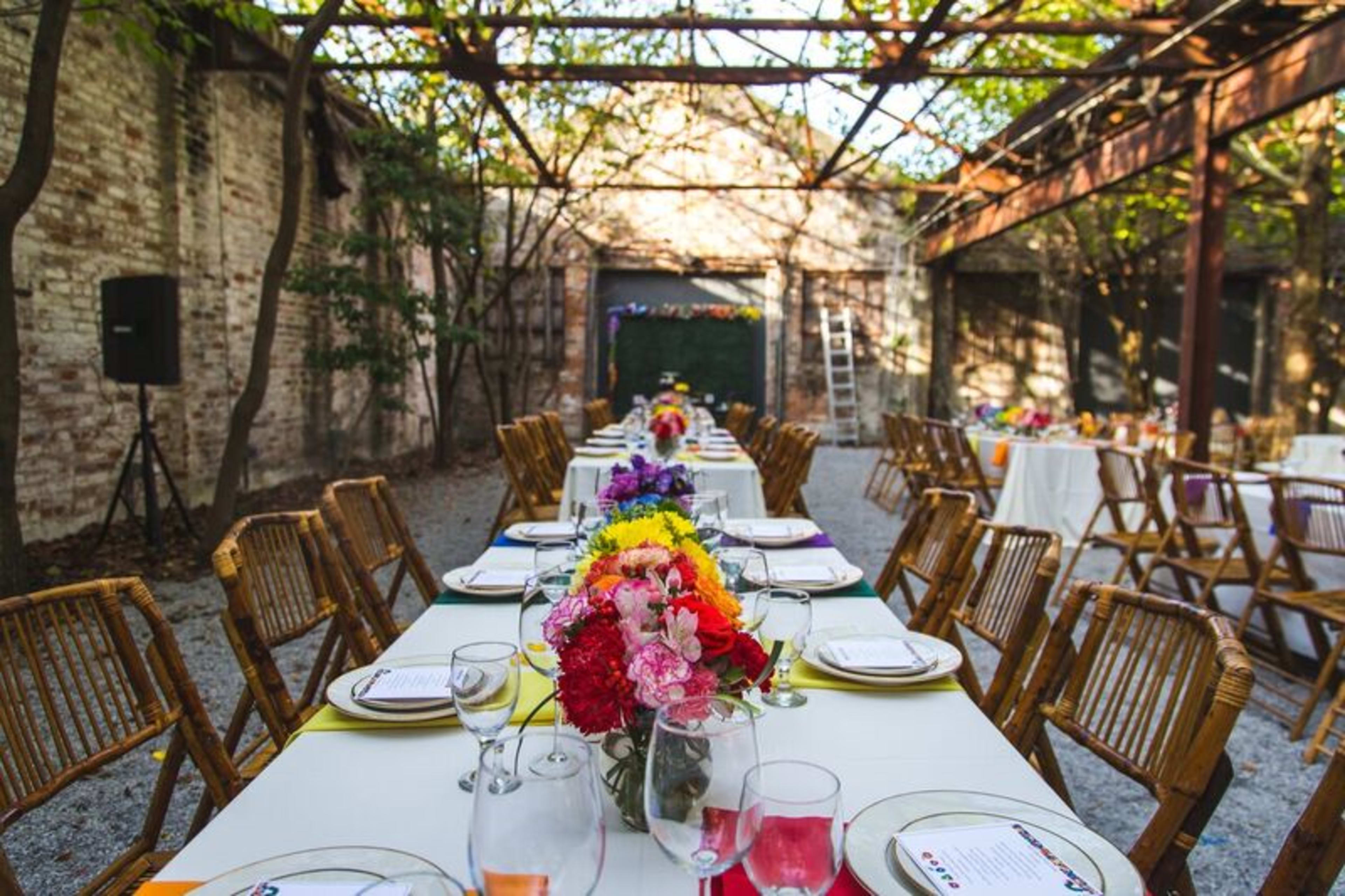 A long table is set outdoors under a metal frame, adorned with colorful flowers and tableware, surrounded by rustic brick walls and trees.
