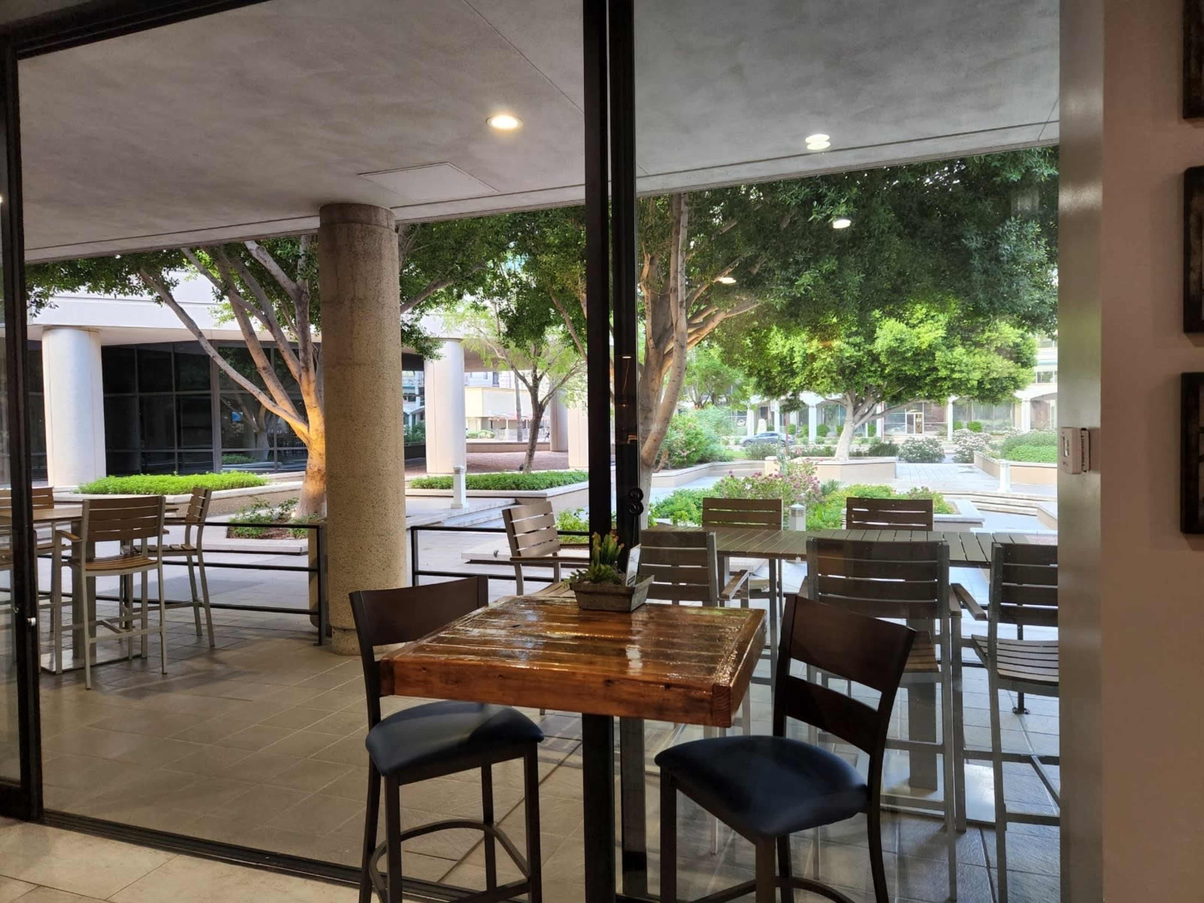 A courtyard view through large glass windows, featuring outdoor seating and trees in a landscaped area.