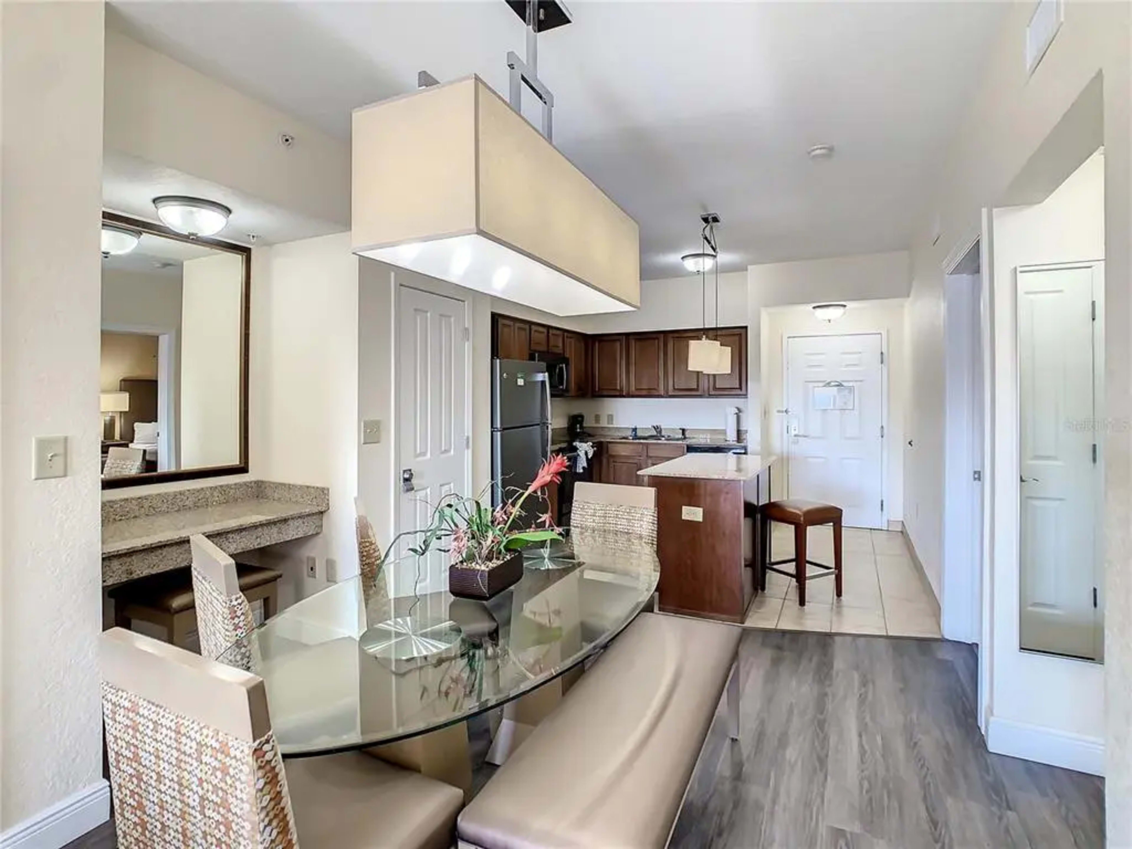 A modern dining area with a glass table, cushioned benches, and a view of an open kitchen featuring wooden cabinets and stainless steel appliances.
