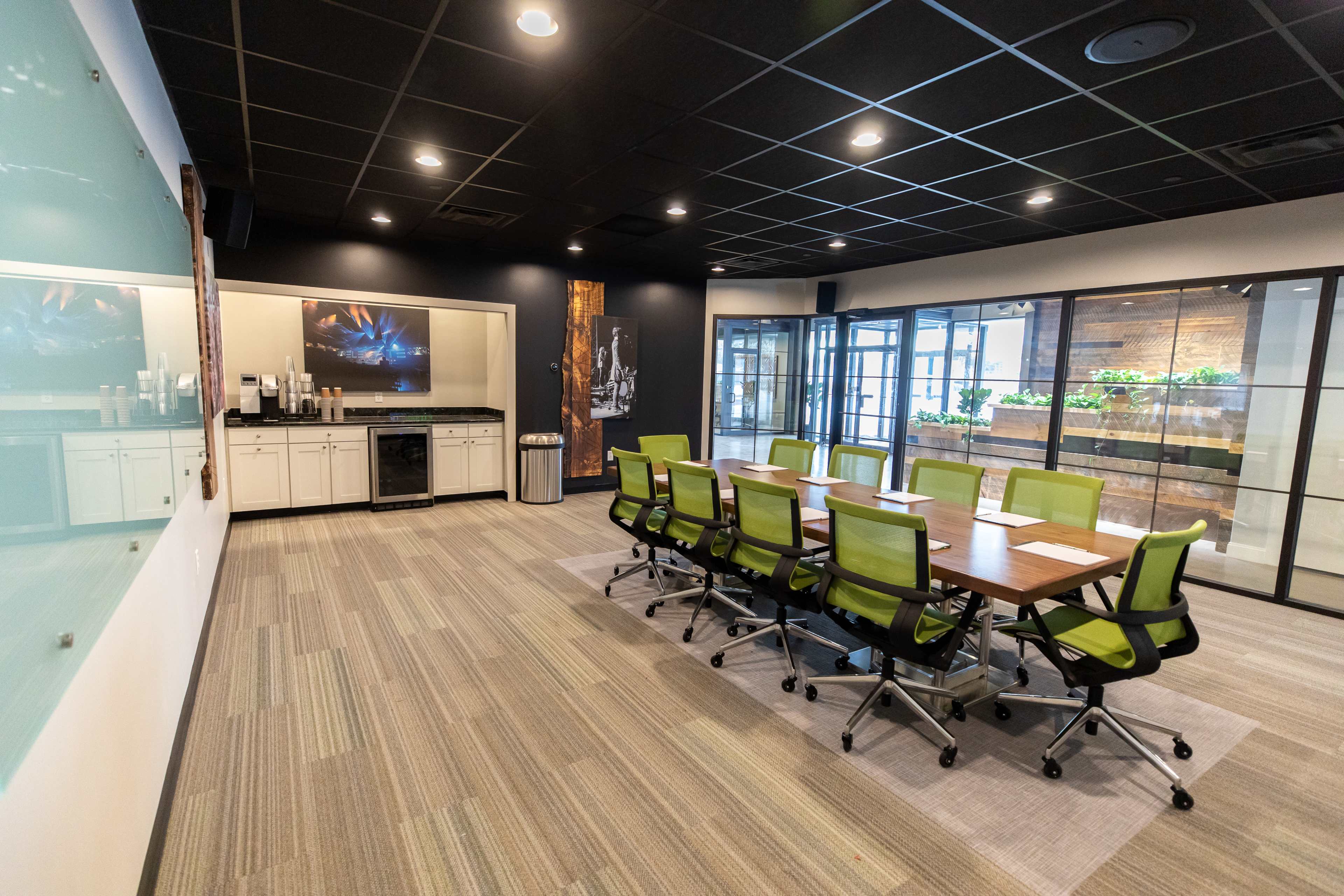 A modern conference room features a long wooden table surrounded by green rolling chairs, with a kitchenette and glass doors leading to a plant-filled area.