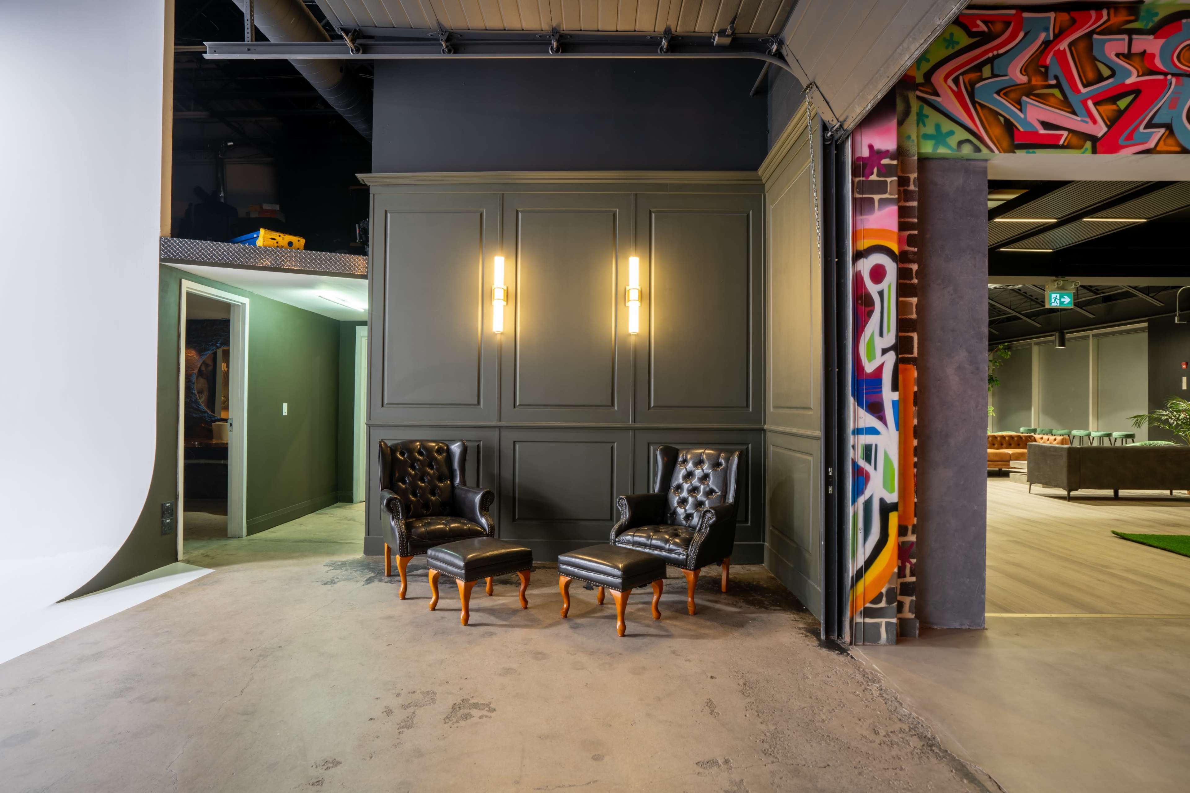 A corner of a modern workspace featuring two black leather chairs with wooden legs against a green wall, next to a brightly colored graffiti mural.