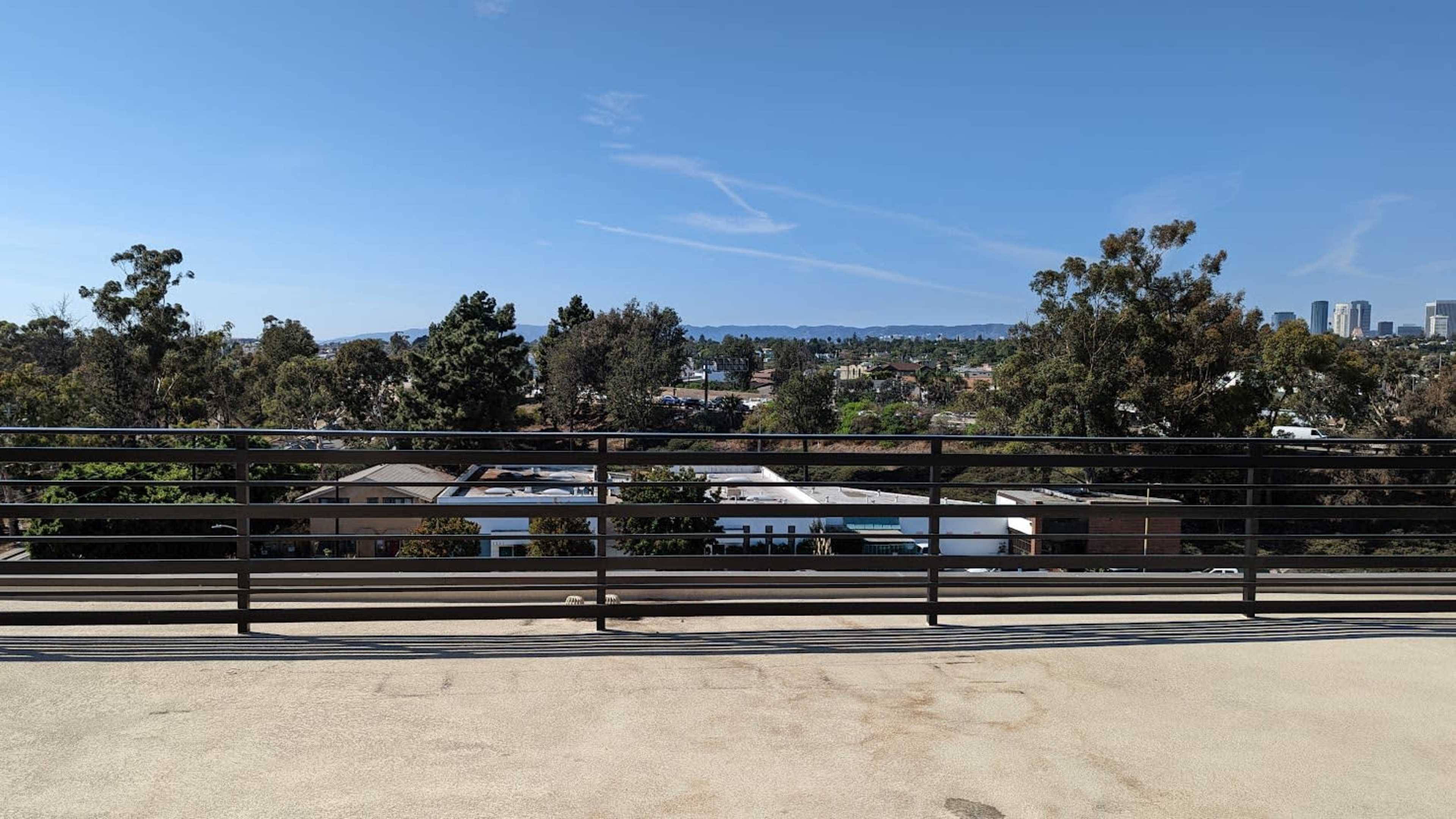 The image shows a railing overlooking a city landscape with buildings and greenery under a clear sky.