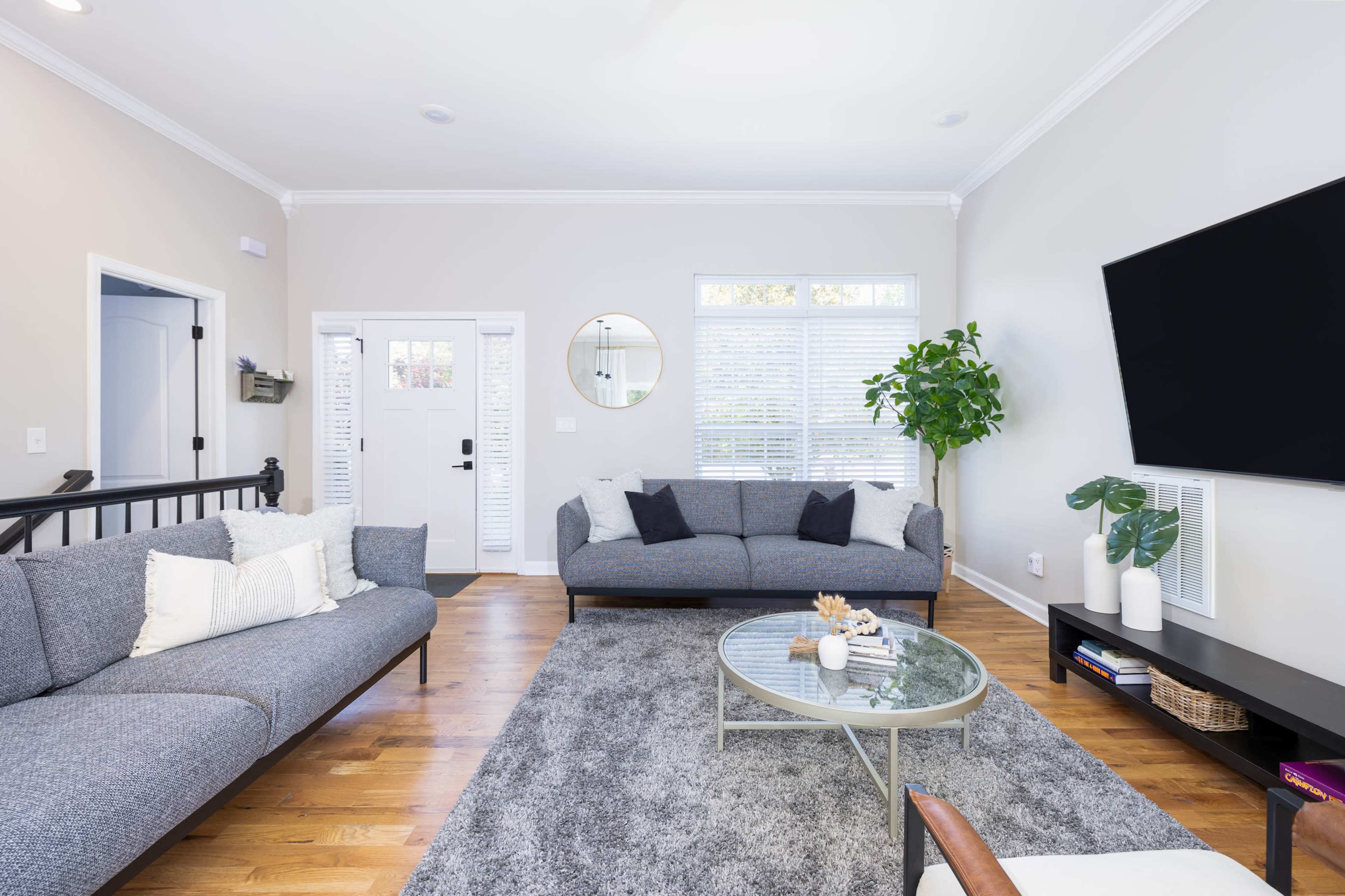A bright living room features two gray sofas, a glass coffee table, and a large wall-mounted television.