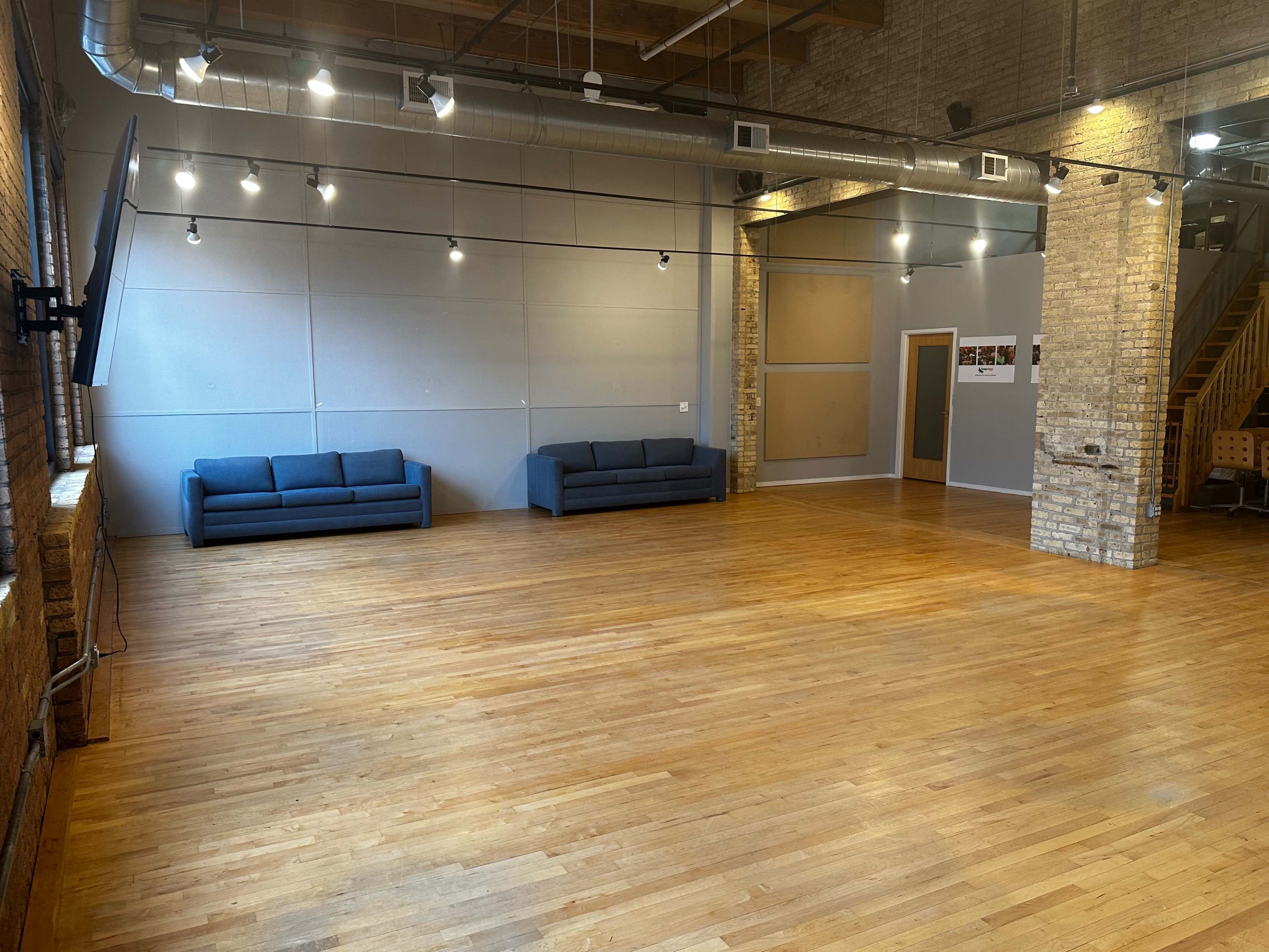 A spacious room with wooden floors, two blue sofas, and exposed brick walls, illuminated by track lighting.