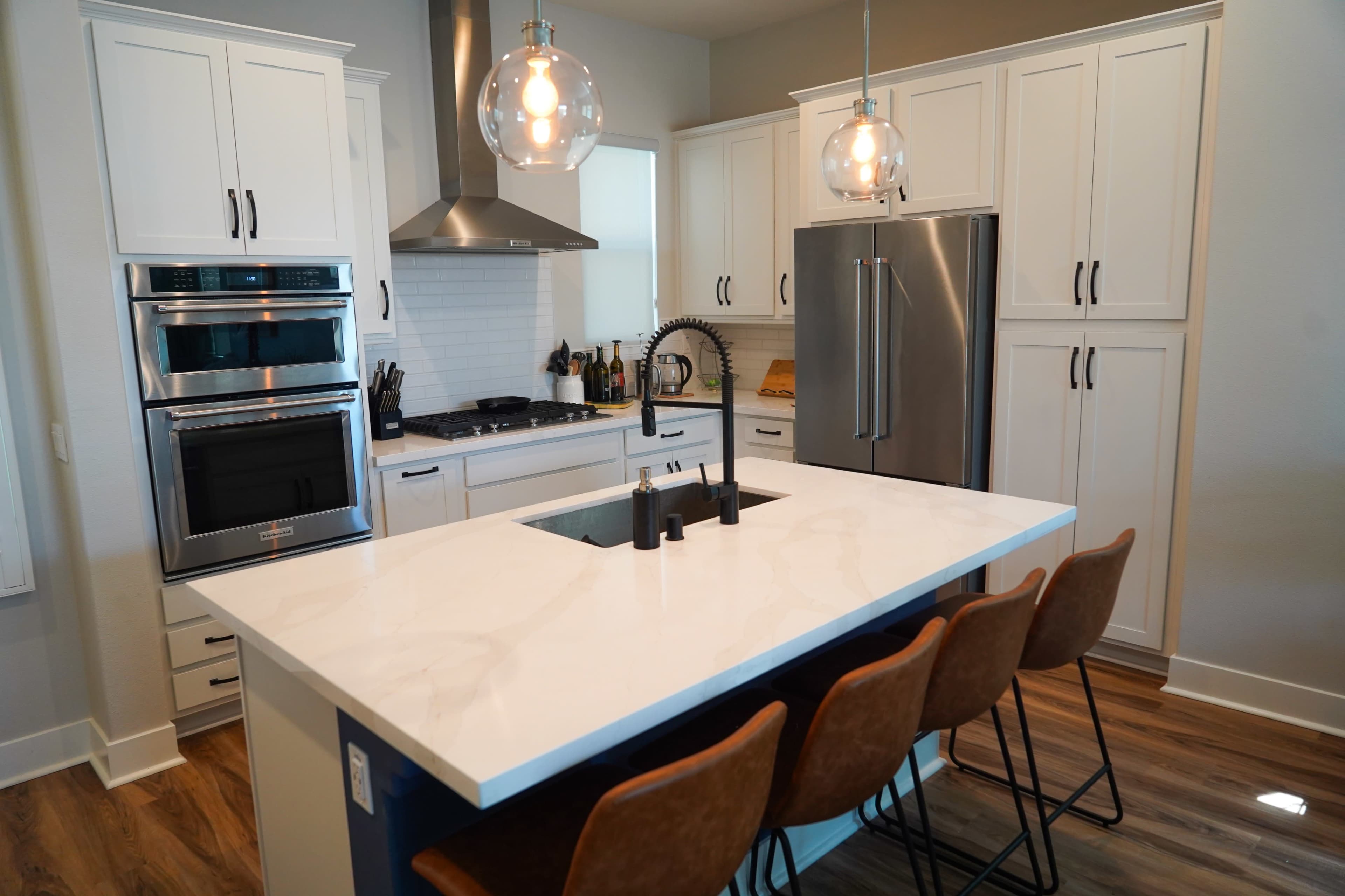 The image shows a modern kitchen with a central island featuring four brown bar stools, stainless steel appliances, and white cabinetry.