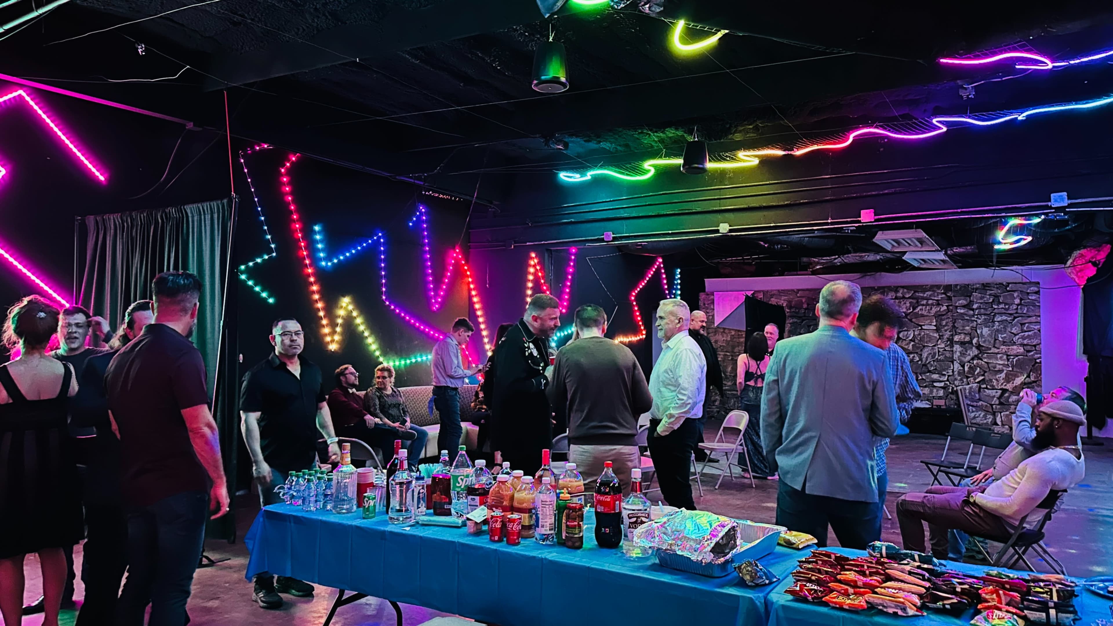 A gathering of people is taking place in a dimly lit room with neon light decorations and a table filled with drinks and snacks.