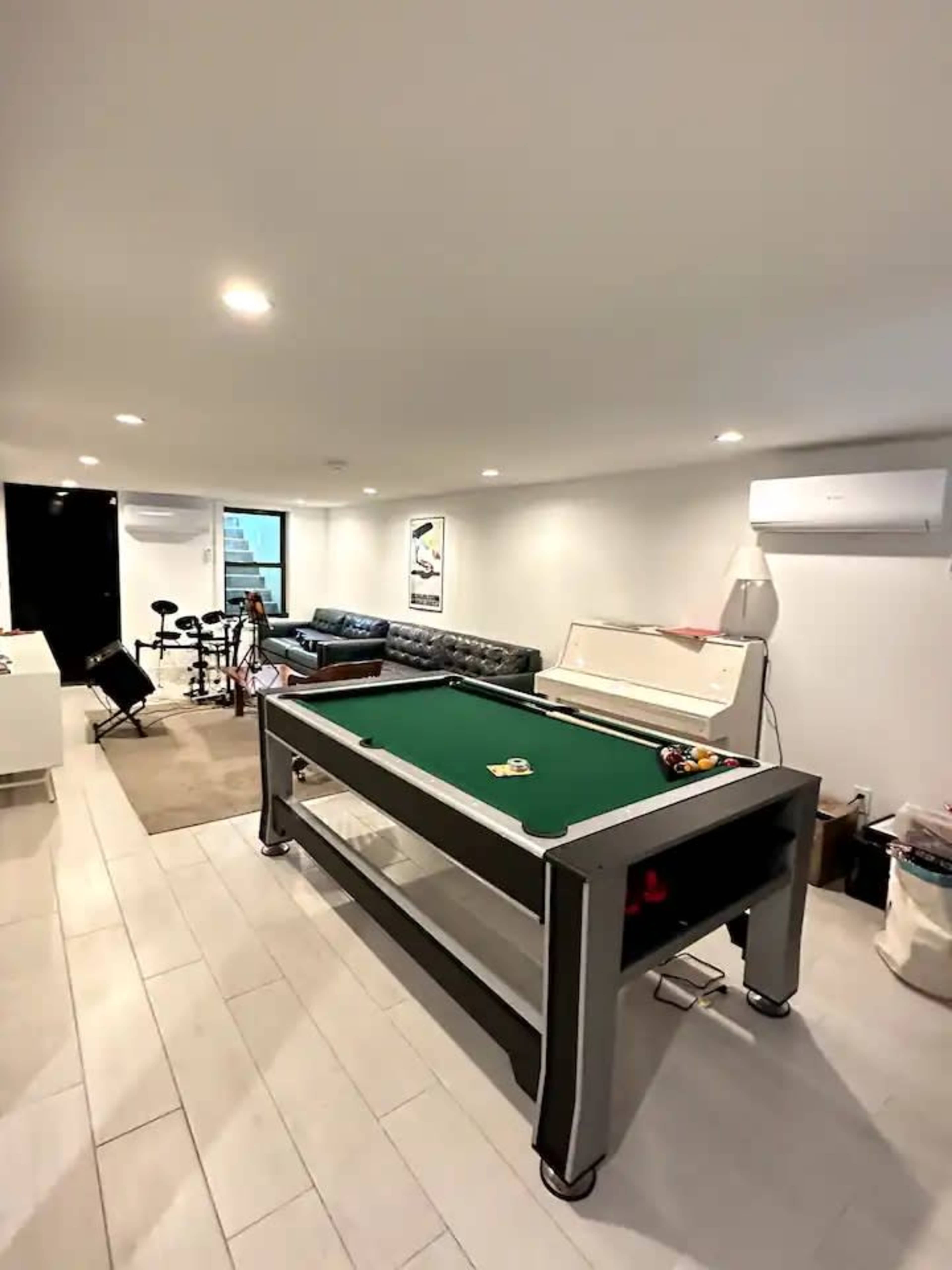 A brightly lit room features a pool table at the center, surrounded by a couch and a set of drums in the corner.