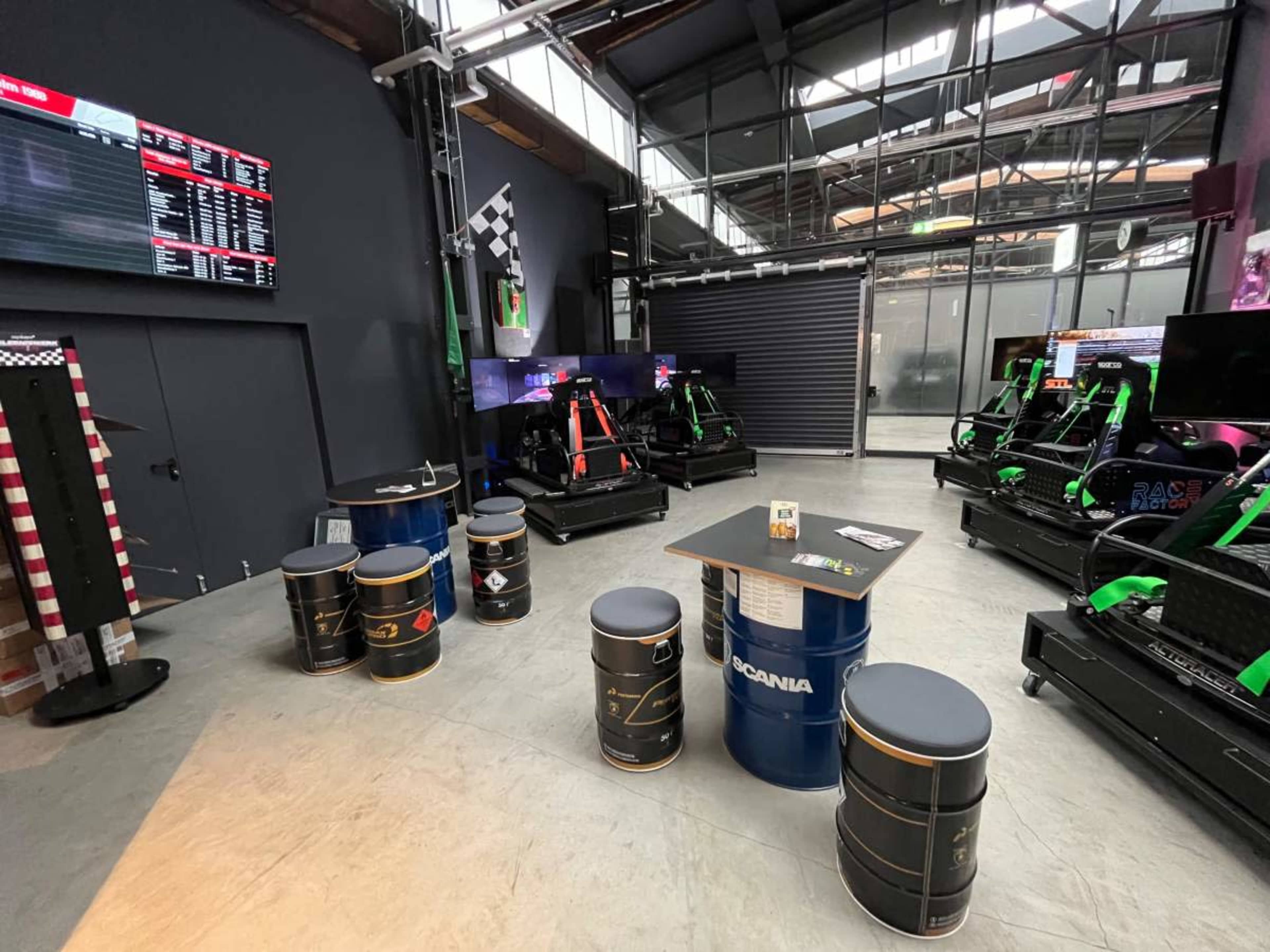 The image shows a racing simulator venue featuring multiple racing rigs, seating made from oil drums, and a large screen displaying race information.