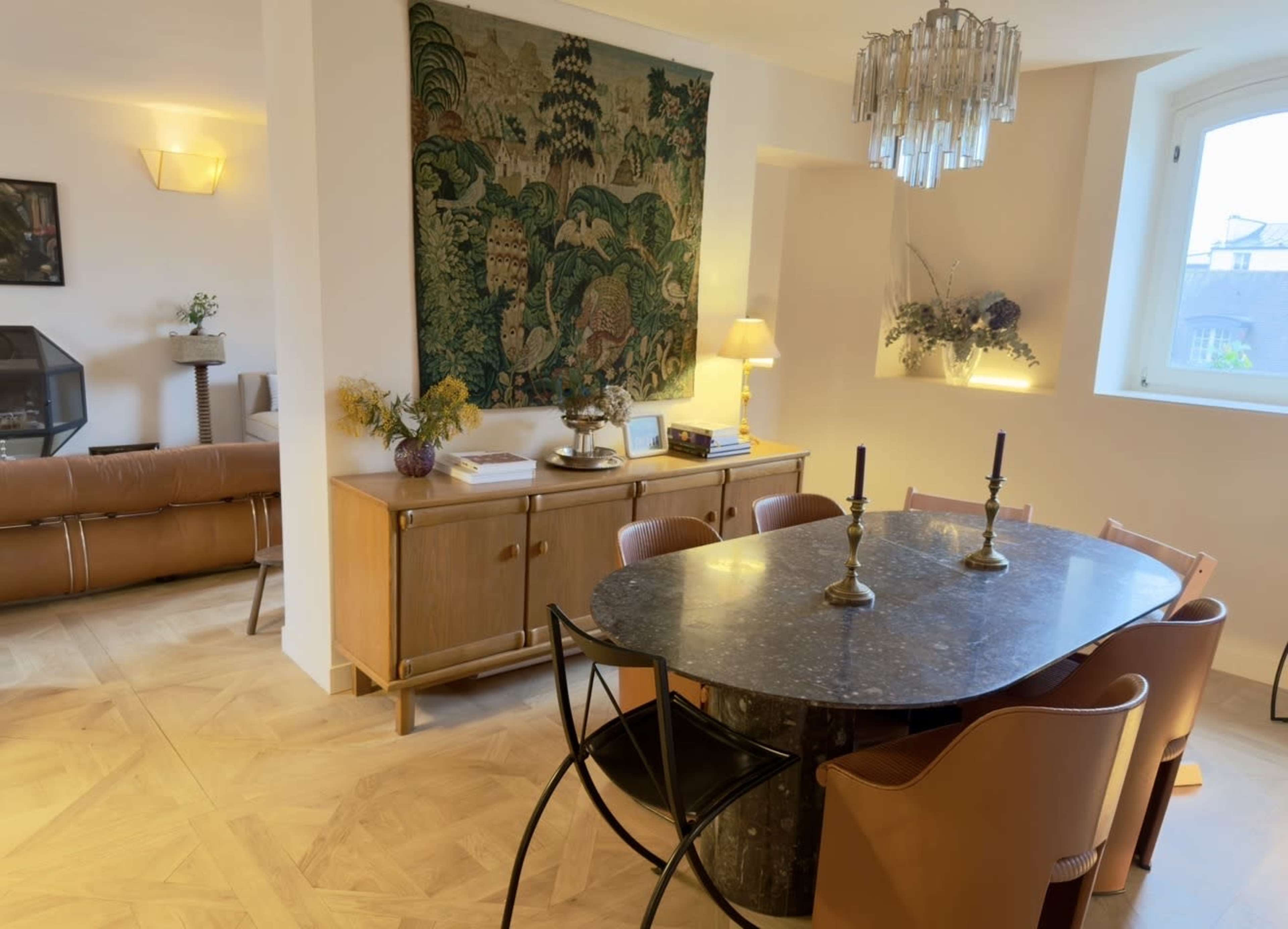 The image shows a dining area with a round marble table surrounded by brown leather chairs, and a large tapestry hanging on the wall above a wooden sideboard.