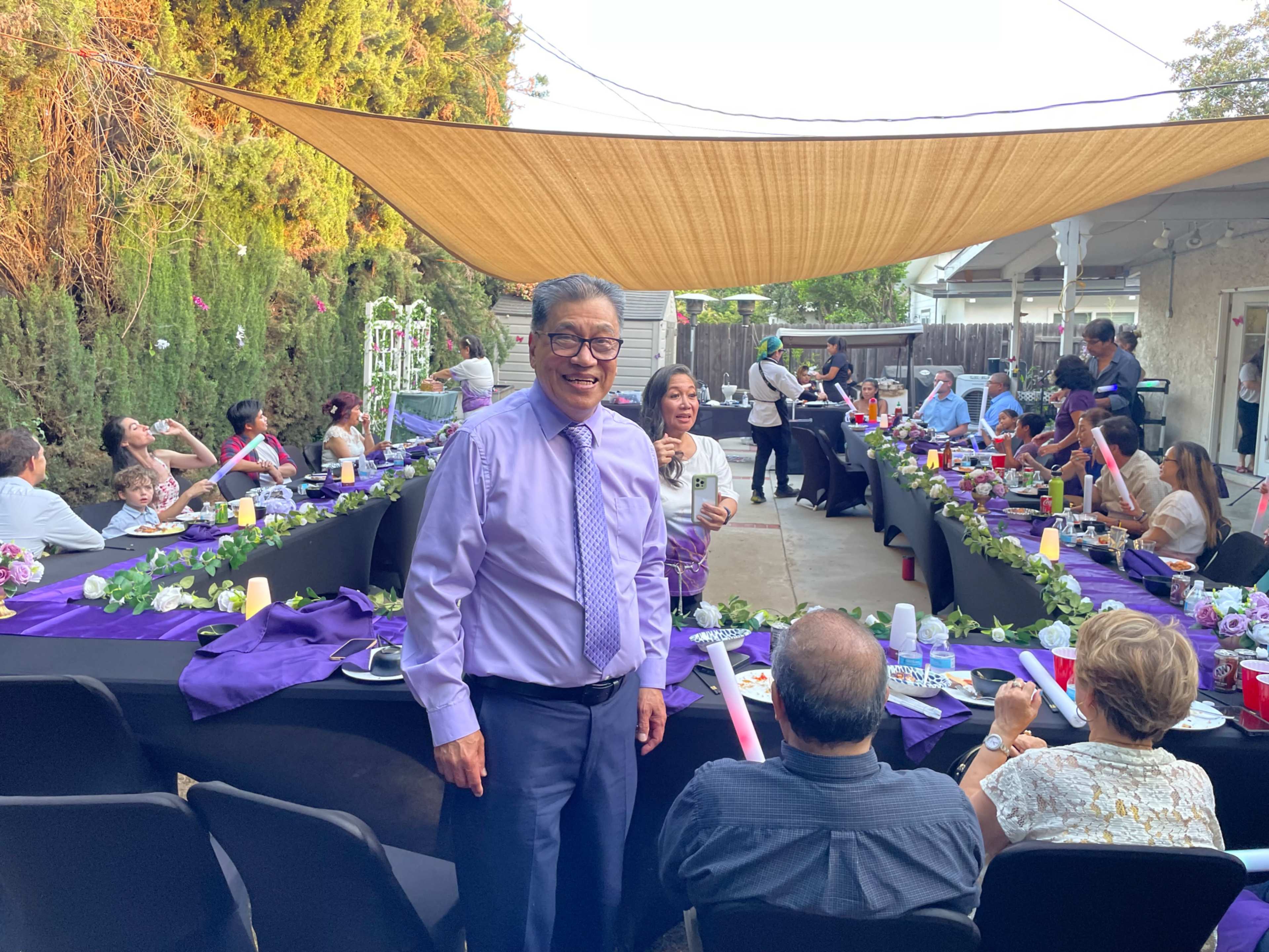 A man in a purple shirt smiles while standing in a backyard filled with guests seated at tables adorned with purple tablecloths and decorations.
