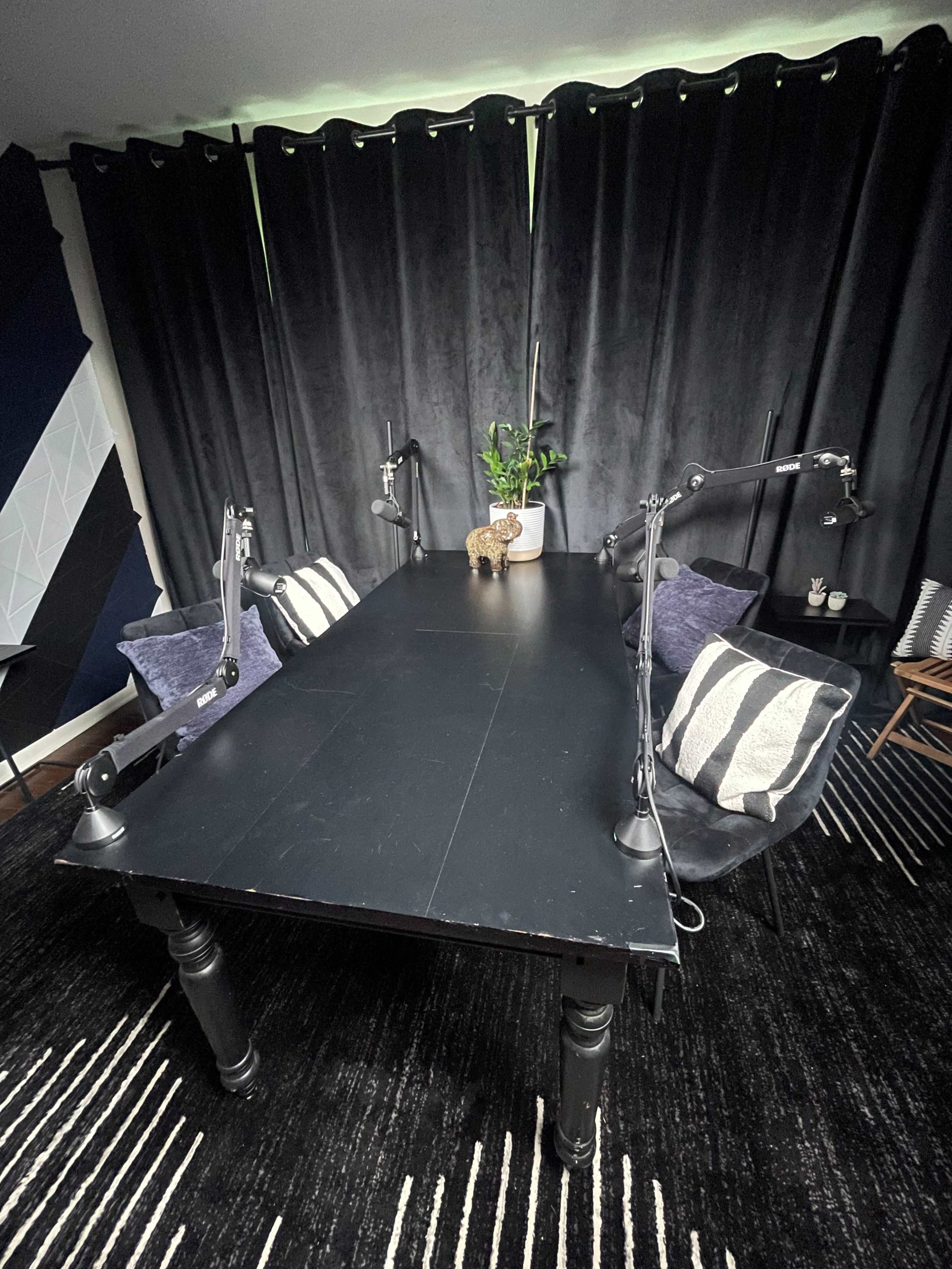 A large black table is positioned in a room with dark curtains, accompanied by two desk lamps and decorative pillows on black chairs.