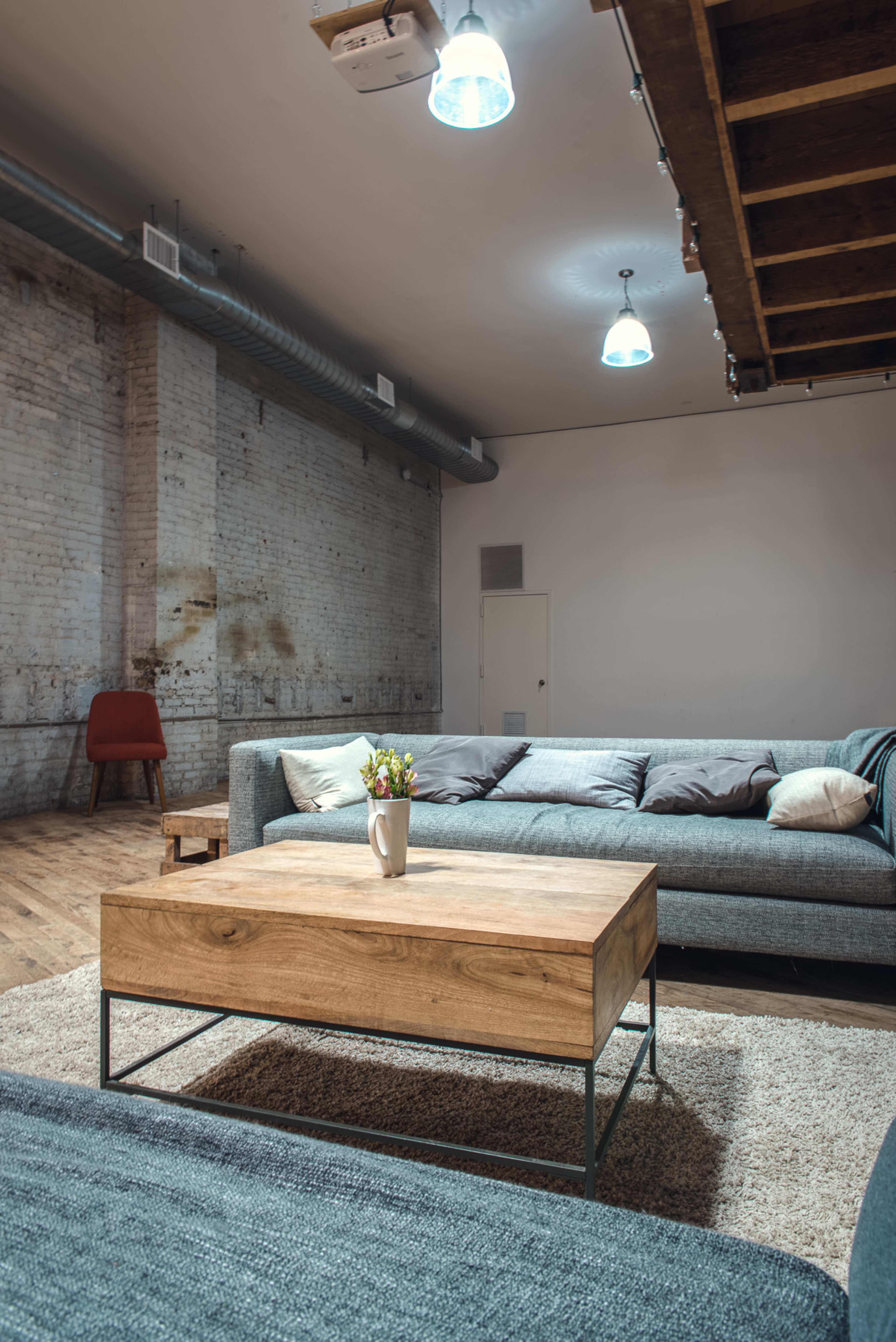 A modern living room features a gray sectional sofa, a wooden coffee table, and a vase of flowers on a light rug, with exposed brick walls and overhead lighting.