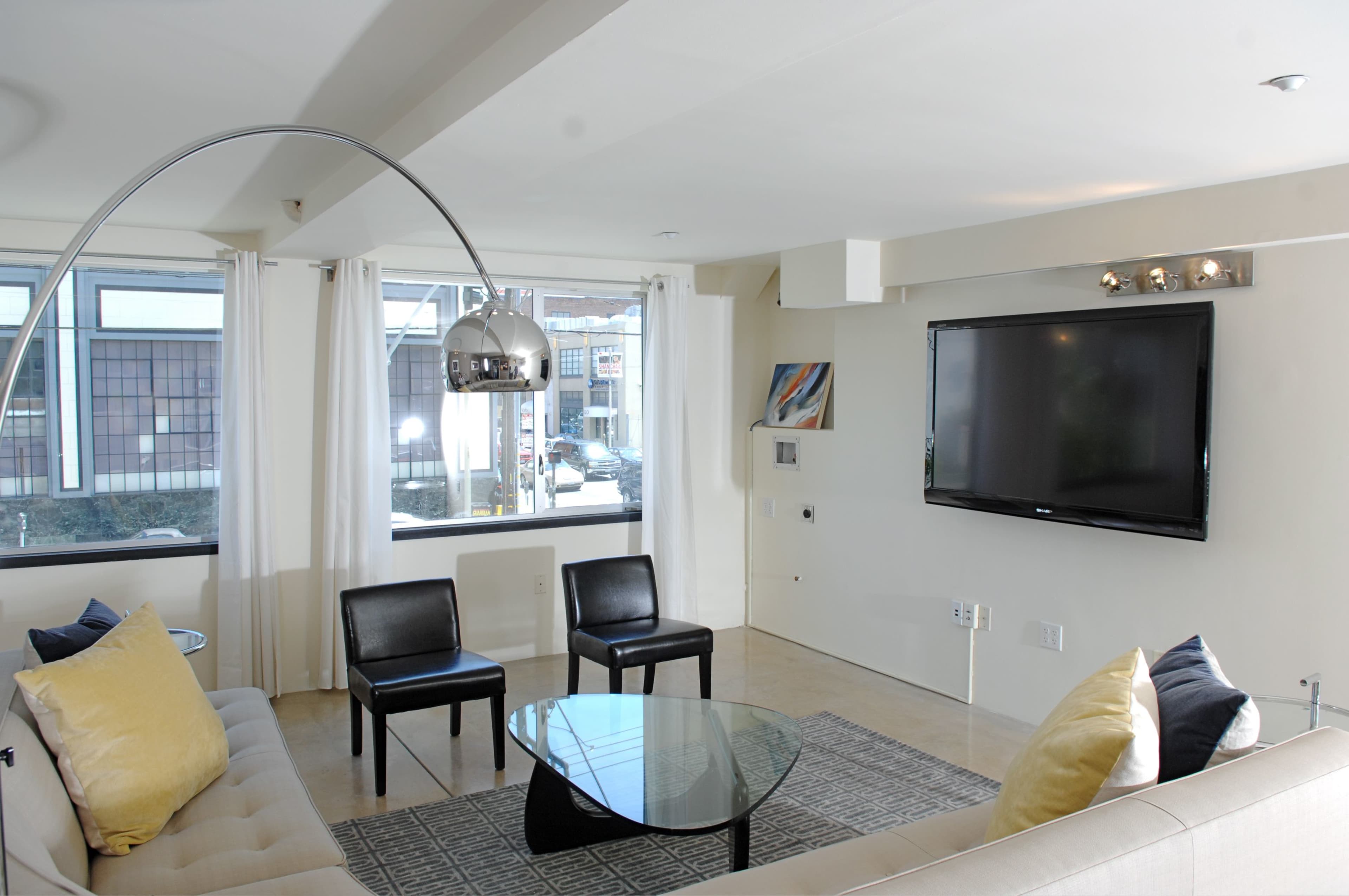 A modern living room with a sofa, two black chairs, a glass coffee table, and a wall-mounted TV.