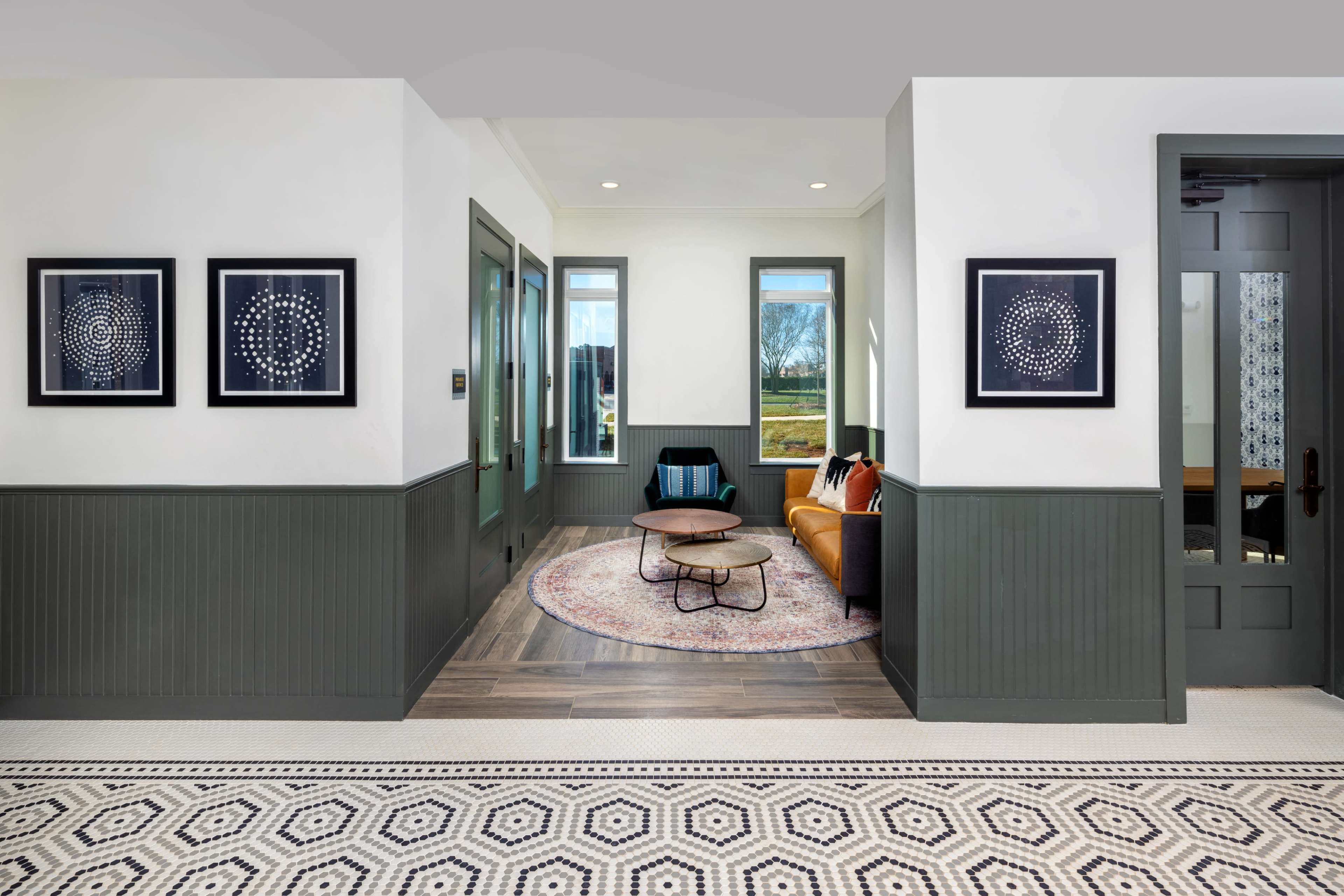 A modern hallway with a cozy seating area featuring a circular rug and a sofa, framed artwork on the walls, and large windows allowing natural light to enter.