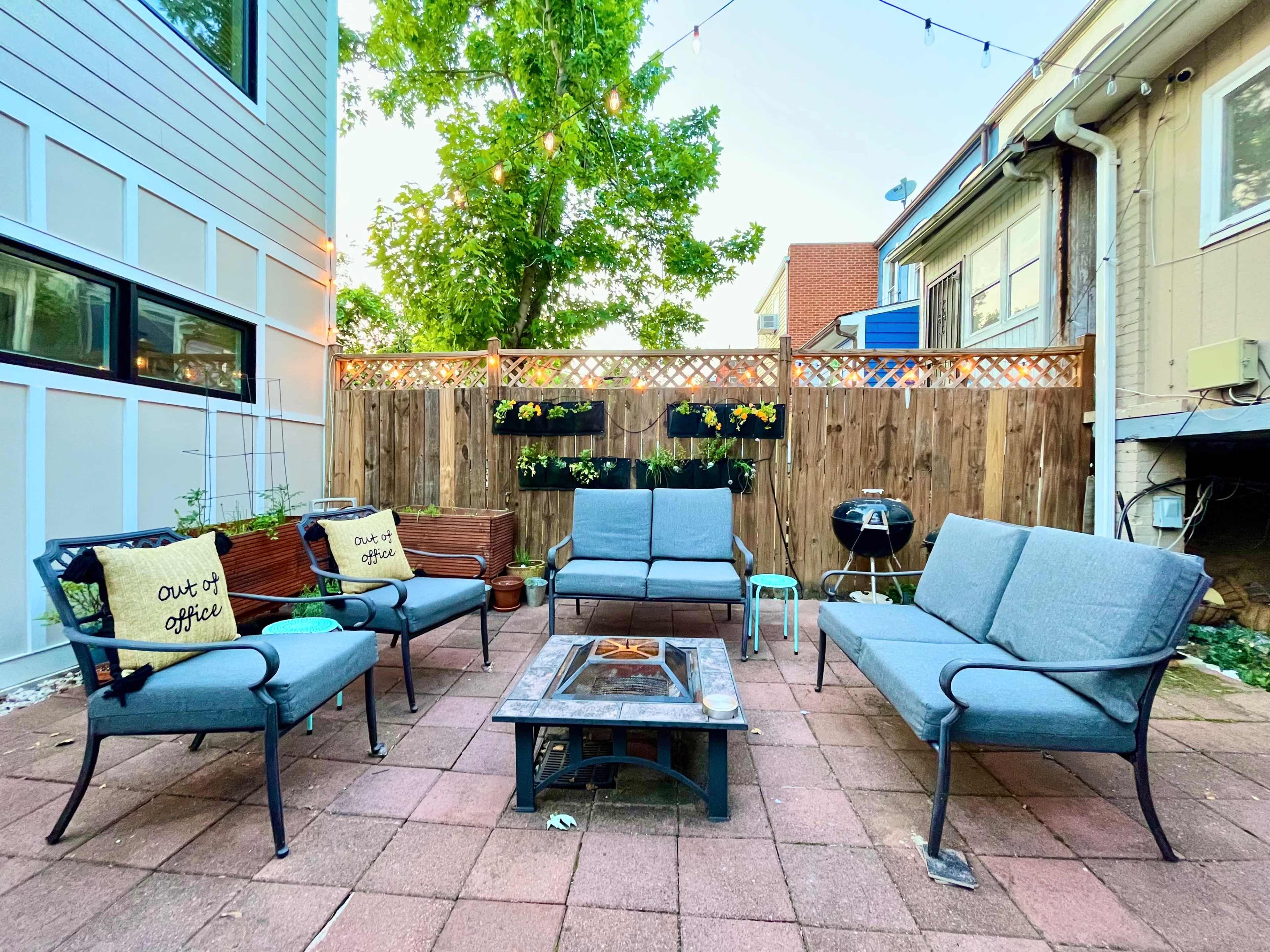 A patio area features a fire pit surrounded by several seating arrangements, planters, and string lights overhead.